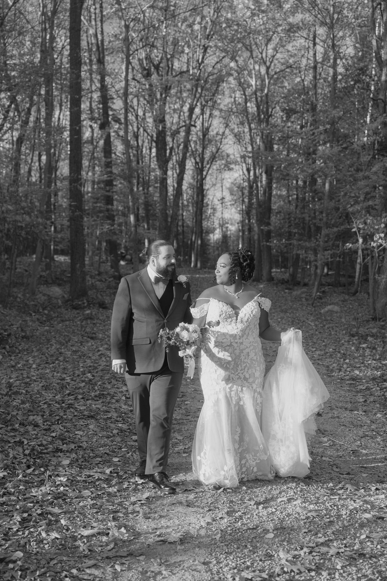 A couple in wedding attire stands together on a forest path covered in fallen leaves during an autumn ceremony.
