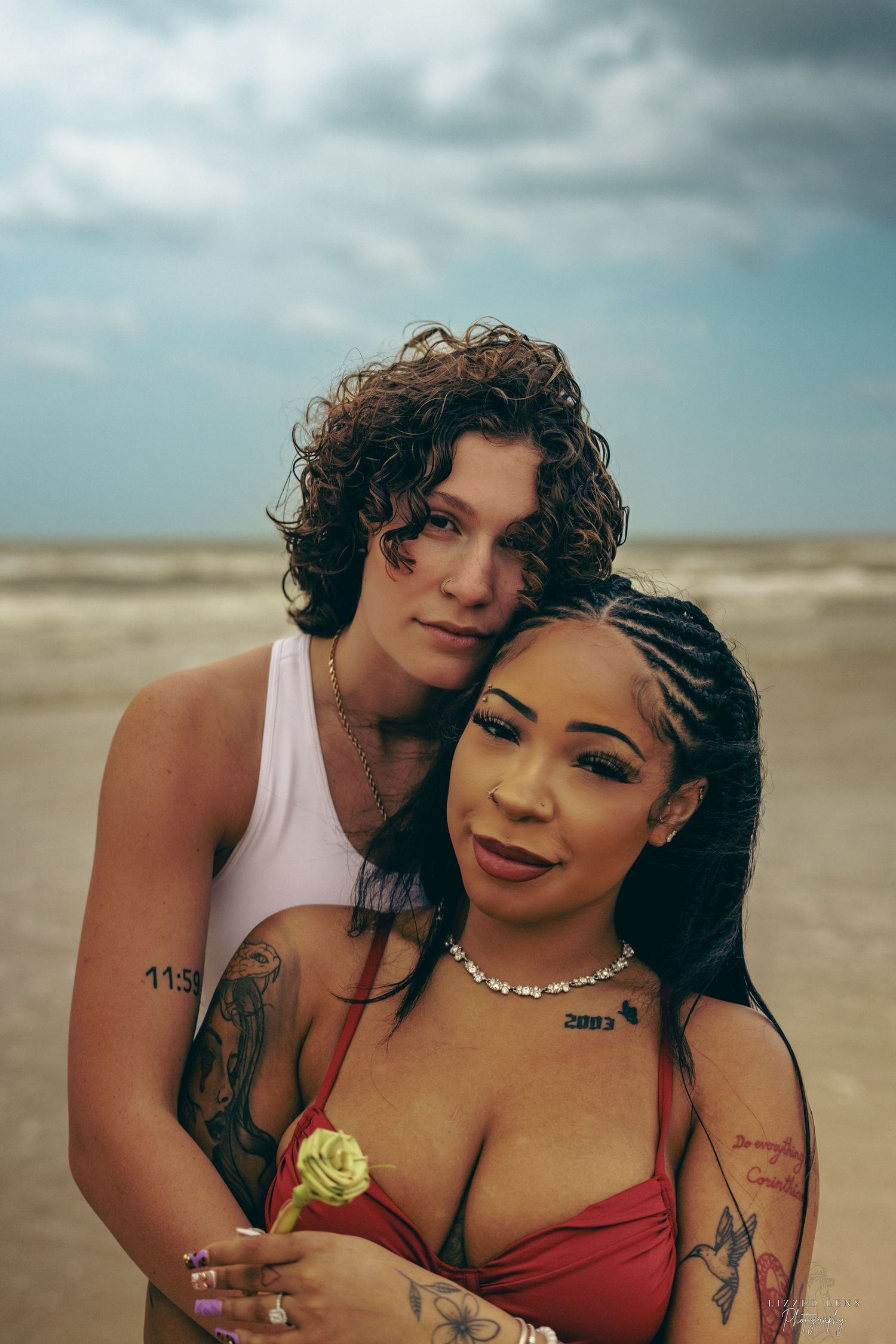 Tattooed couple embracing on a cloudy beach, one holding a yellow rose.