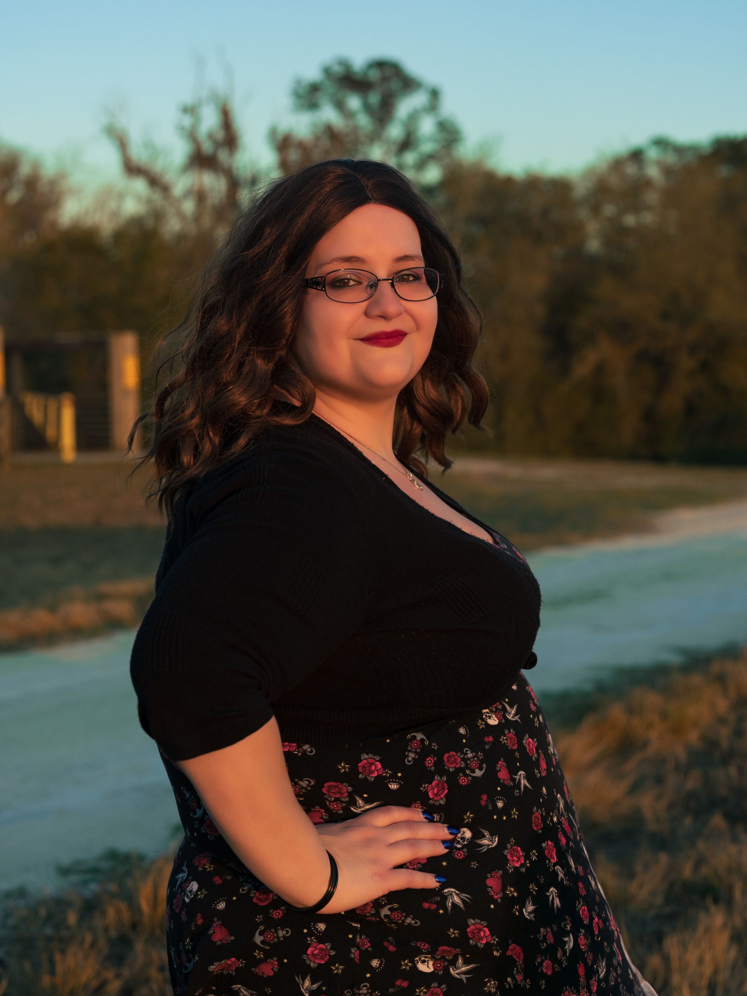 Woman in black top and floral skirt poses outdoors at golden hour with trees in background.