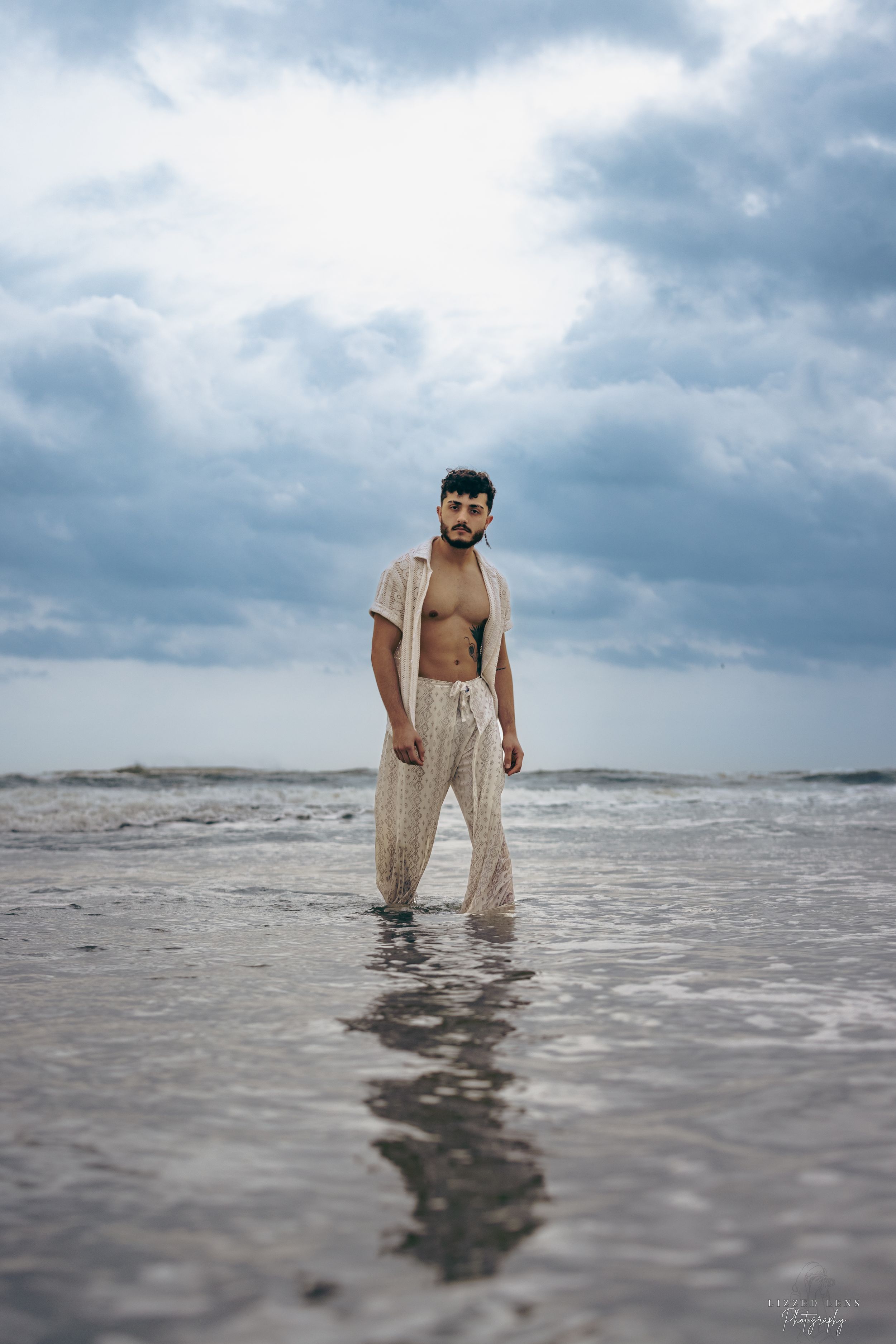Shirtless man in open white shirt standing in shallow ocean water under cloudy sky.