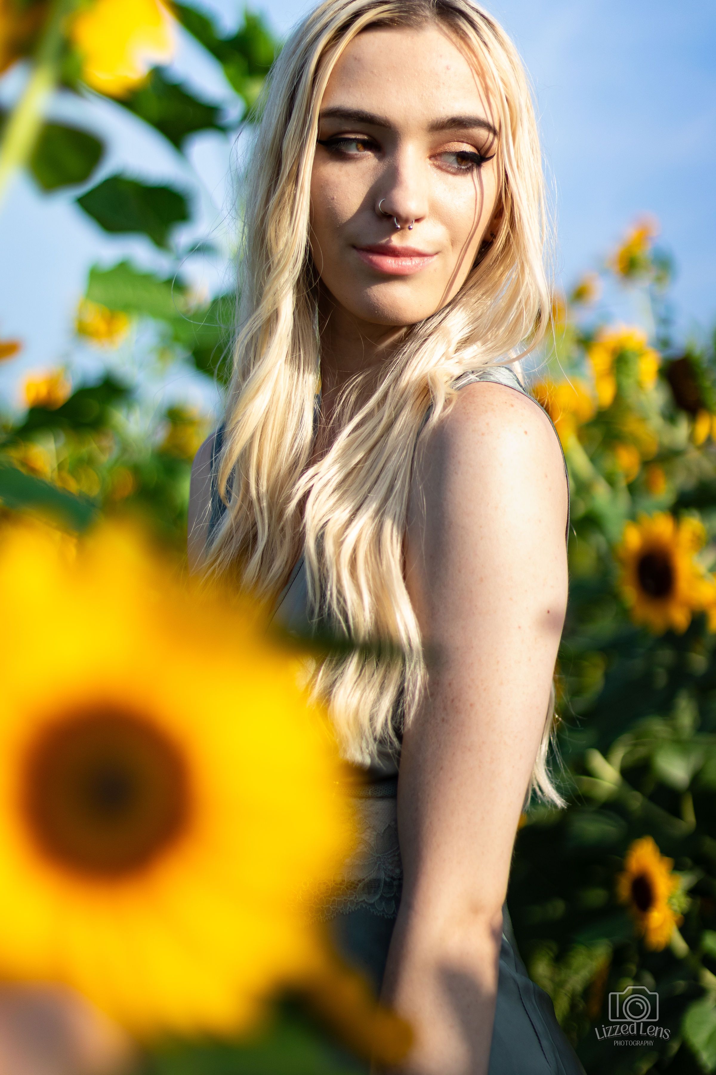 Blonde woman with nose ring standing among sunflowers in golden hour light.