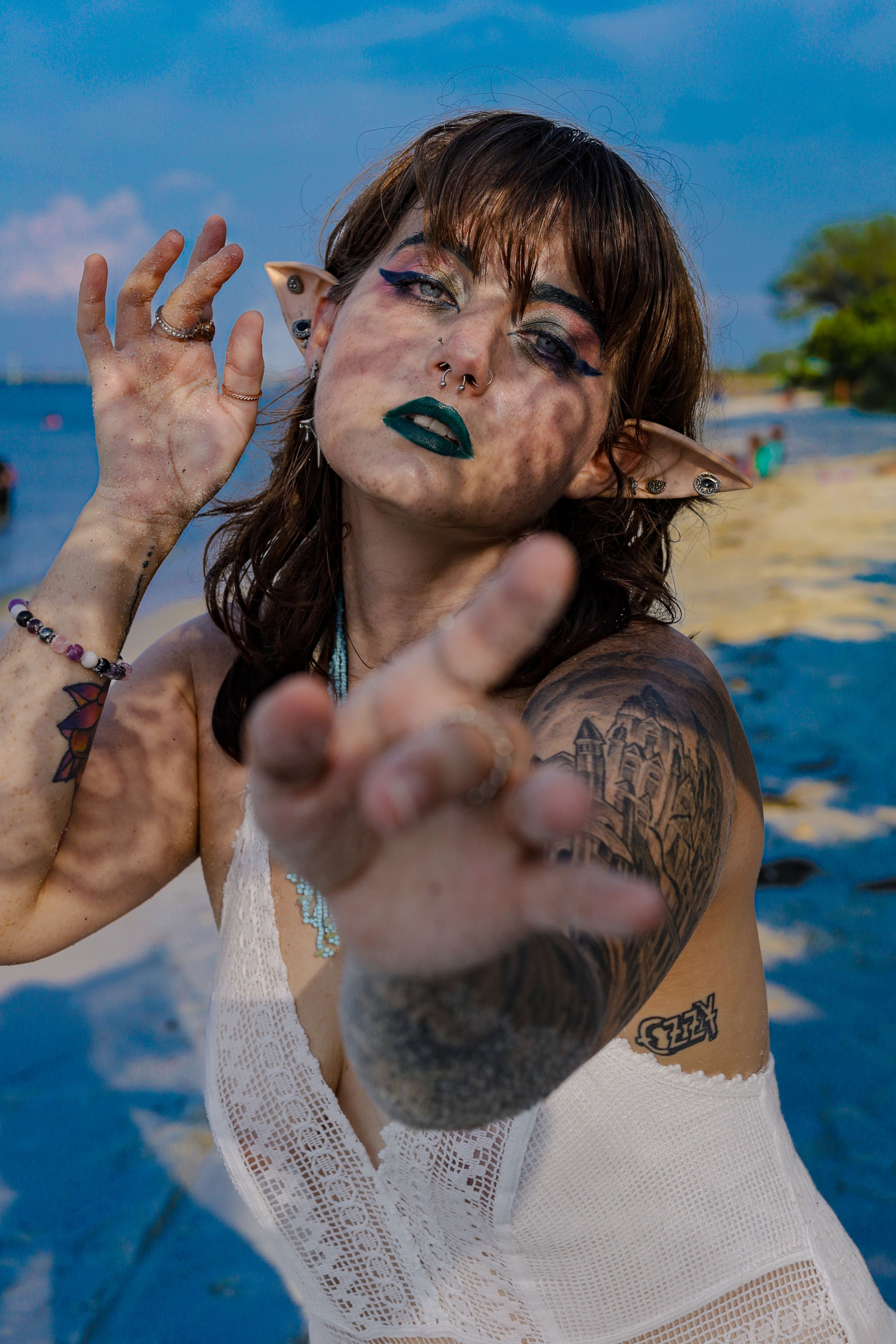 Tattooed woman with green makeup and elf ears reaching toward camera at beach.