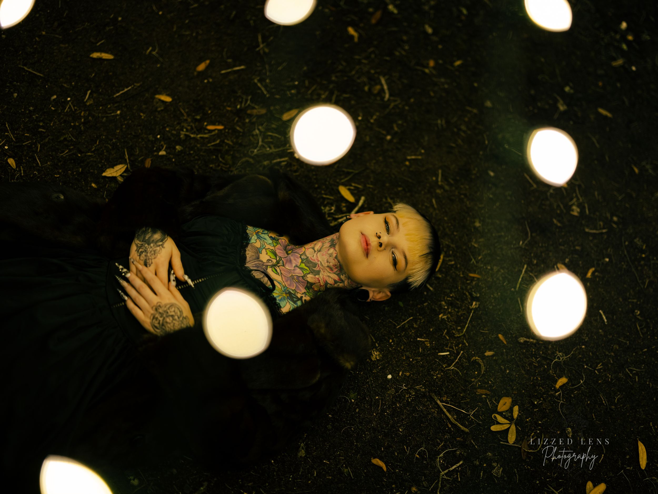 Tattooed person lies on dark ground surrounded by glowing orbs of light in artistic photo.