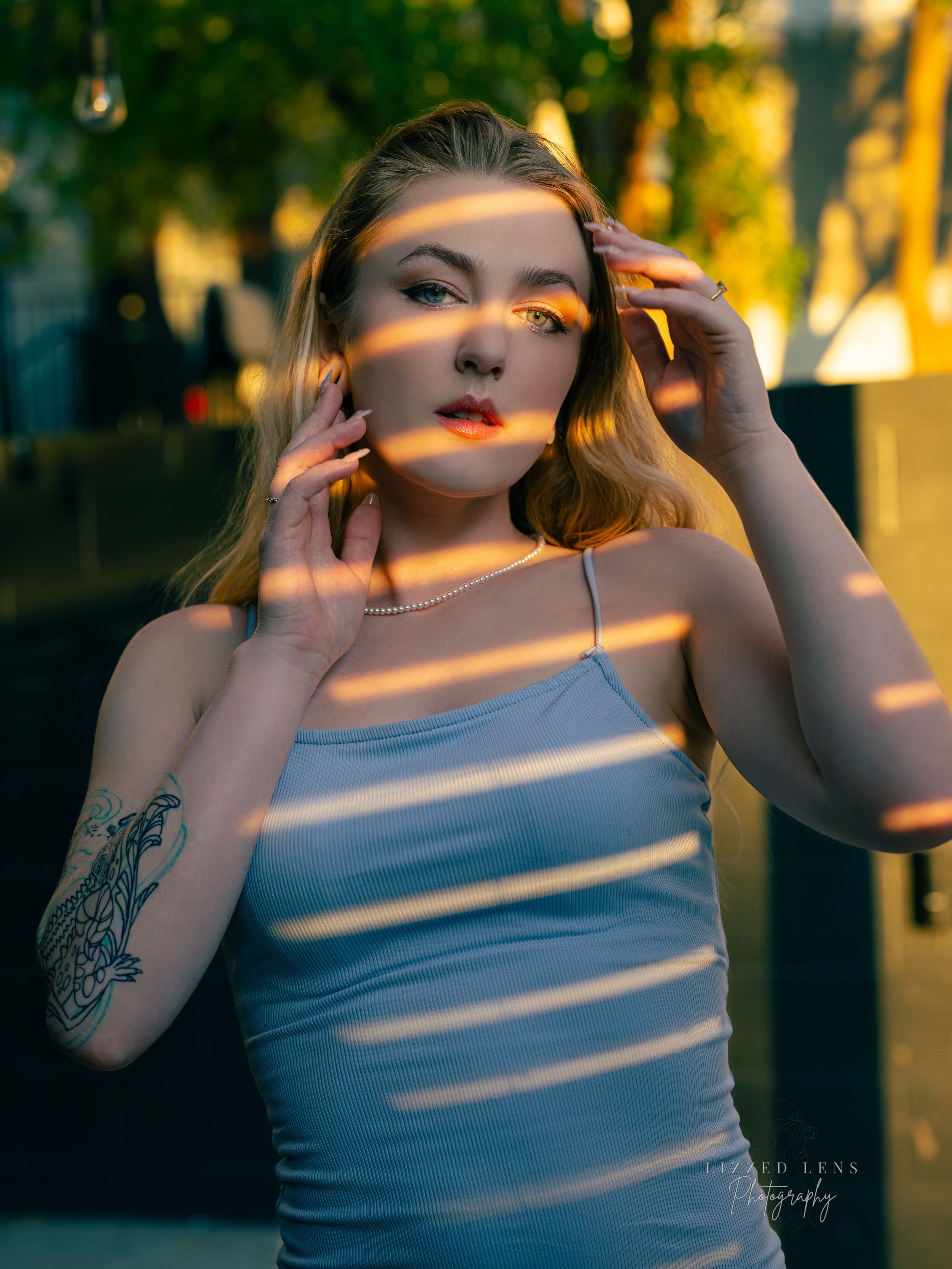 Blonde woman in white tank top with tattoo poses in golden hour light with stripe shadows.