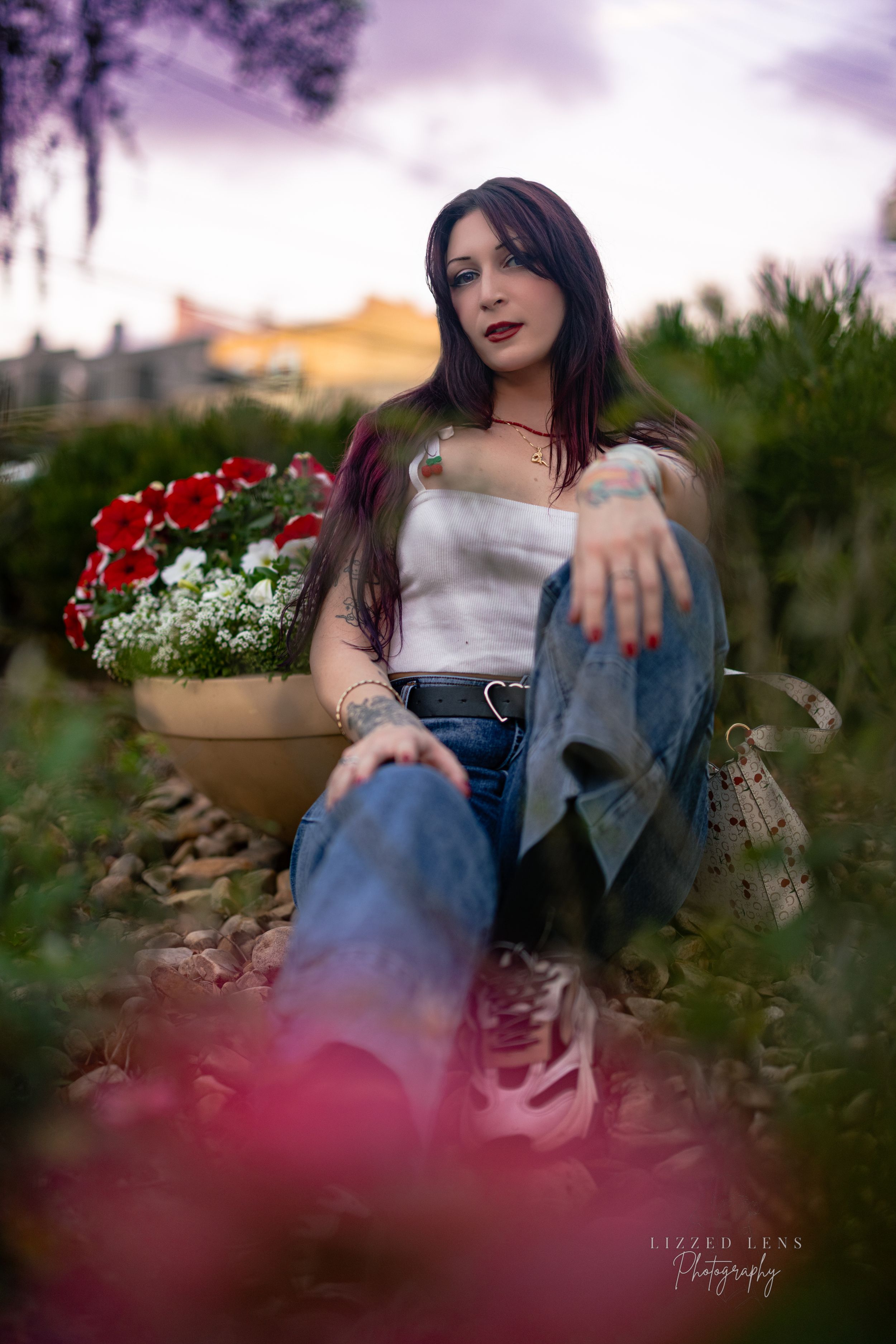 Woman with purple hair sits among garden flowers in jeans at dusk with dramatic sky.