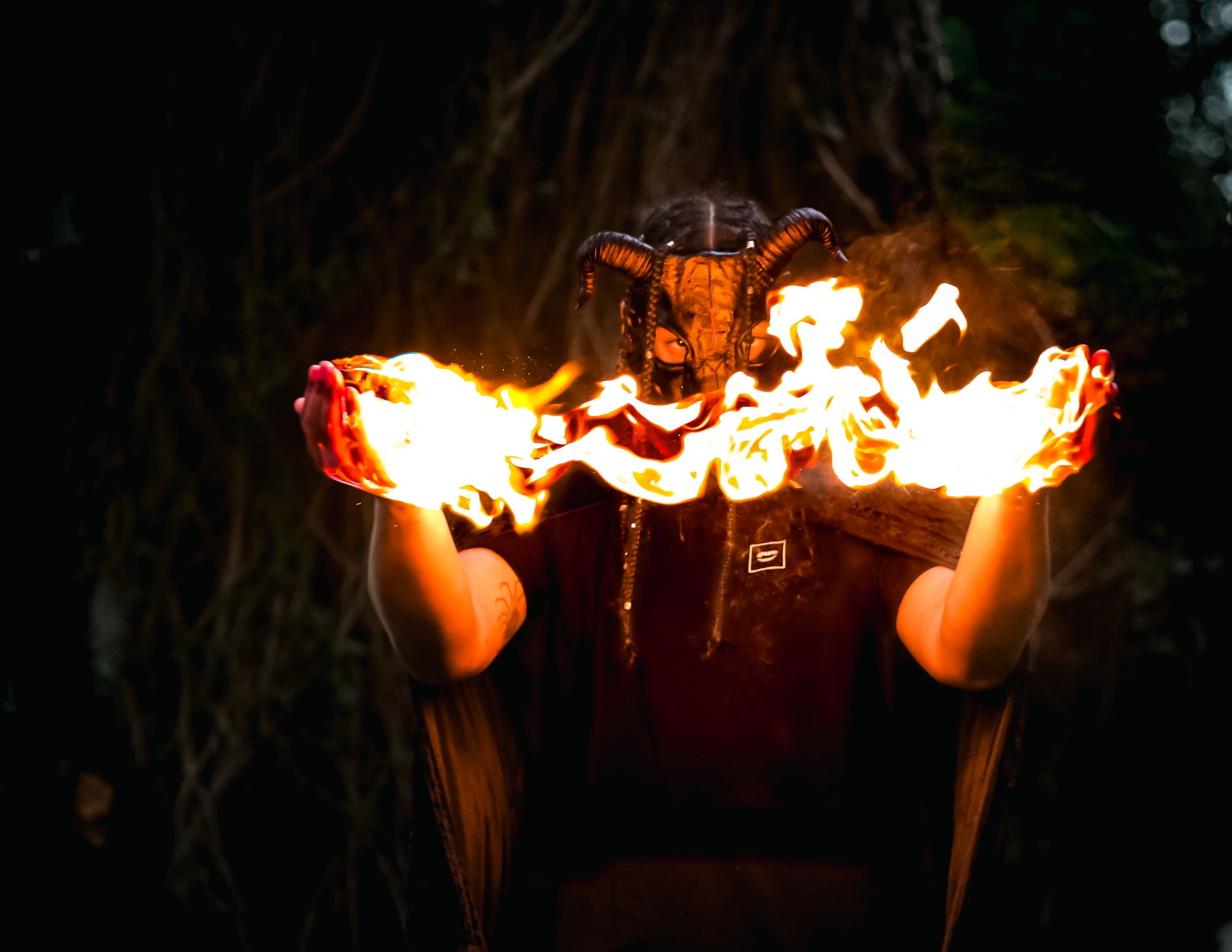 Person wearing horned demon mask holds two balls of fire in dramatic dark forest setting.
