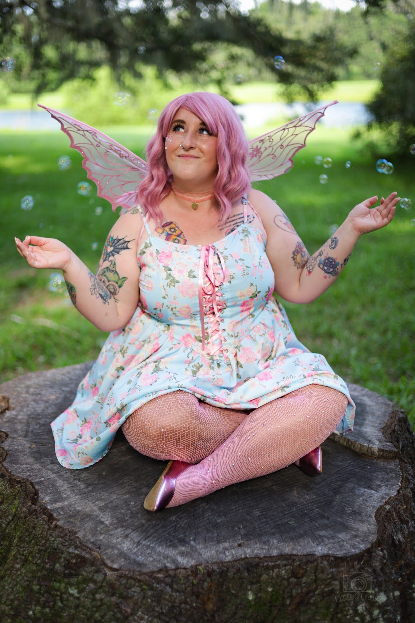 Pink-haired woman in fairy costume with wings sits on tree stump playing with bubbles outdoors.