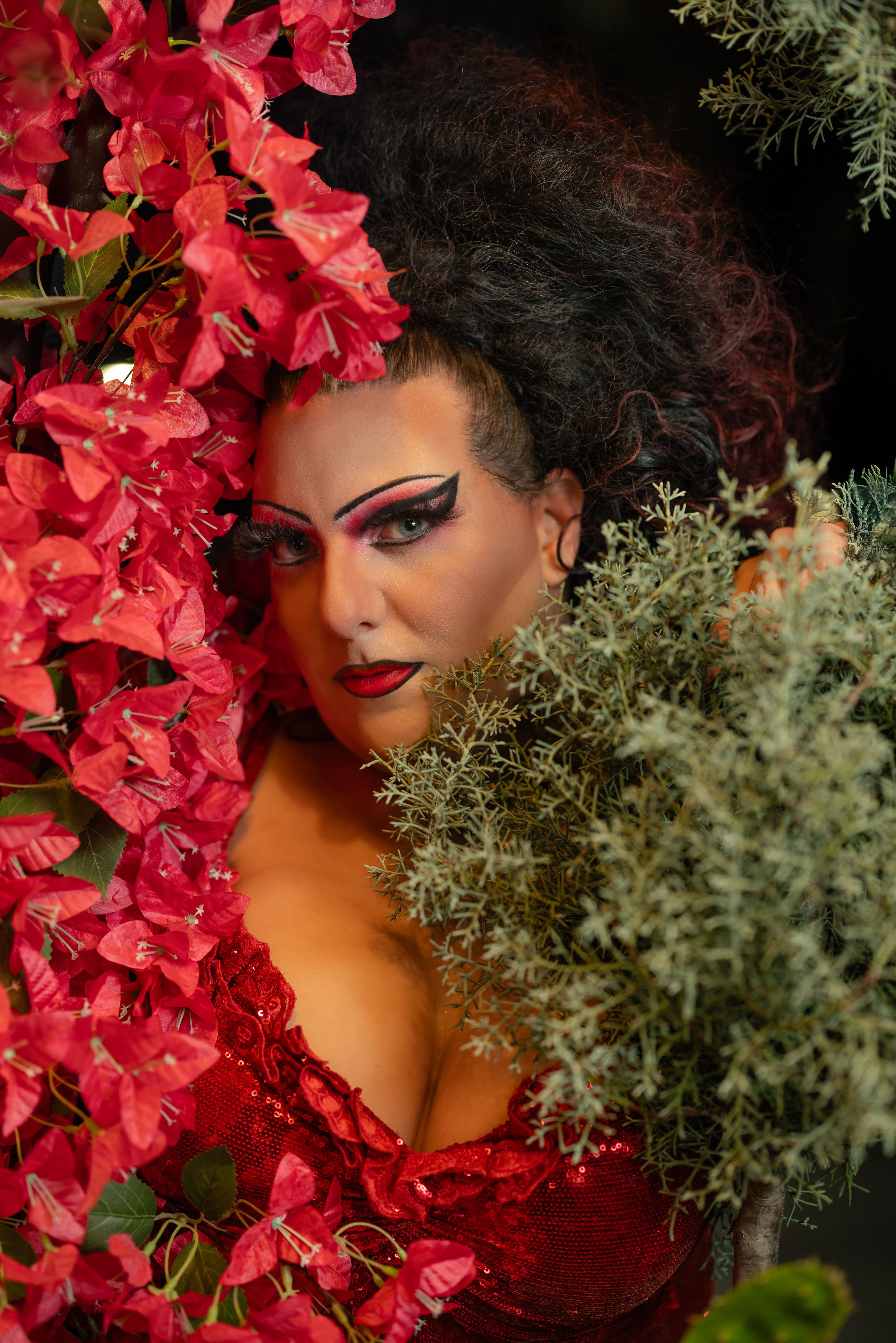 Glamorous woman with dramatic makeup surrounded by vibrant red bougainvillea flowers and green foliage.