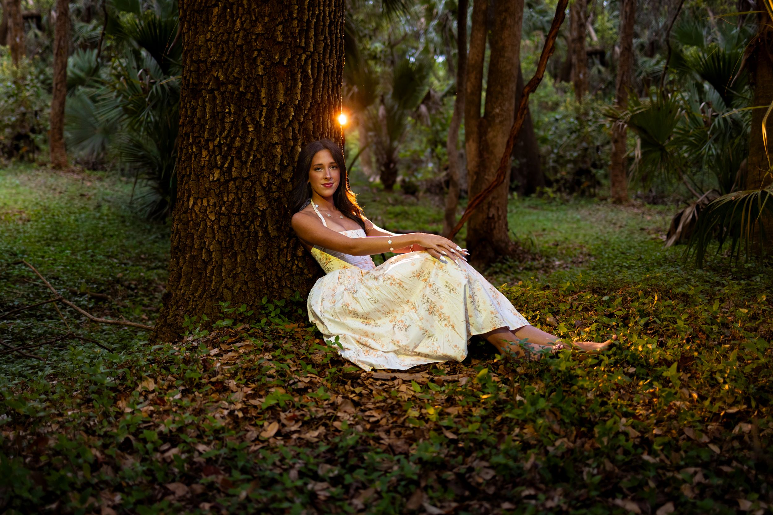 Woman in yellow floral dress sitting against a tree in a forest with a glowing light above her head.