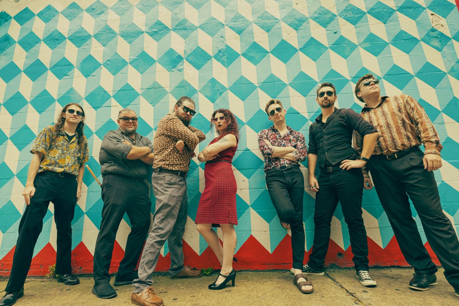 Seven-member band poses casually against a vibrant teal and white geometric patterned wall in an outdoor setting.