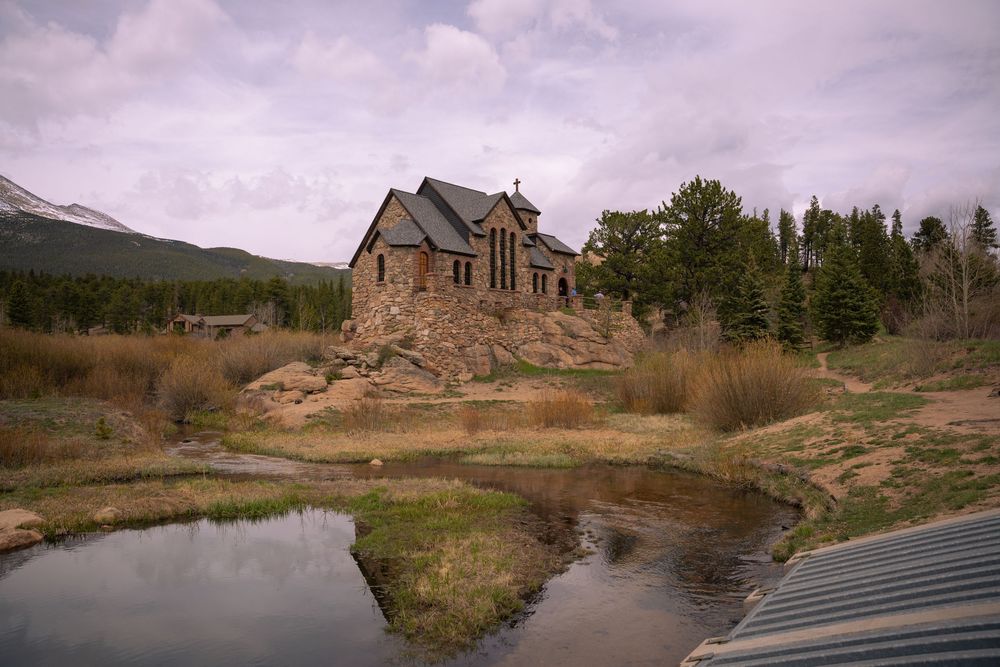 A panoramic view of a historic stone church near a lake in Colorado with mountains and autumn foliage in the background.