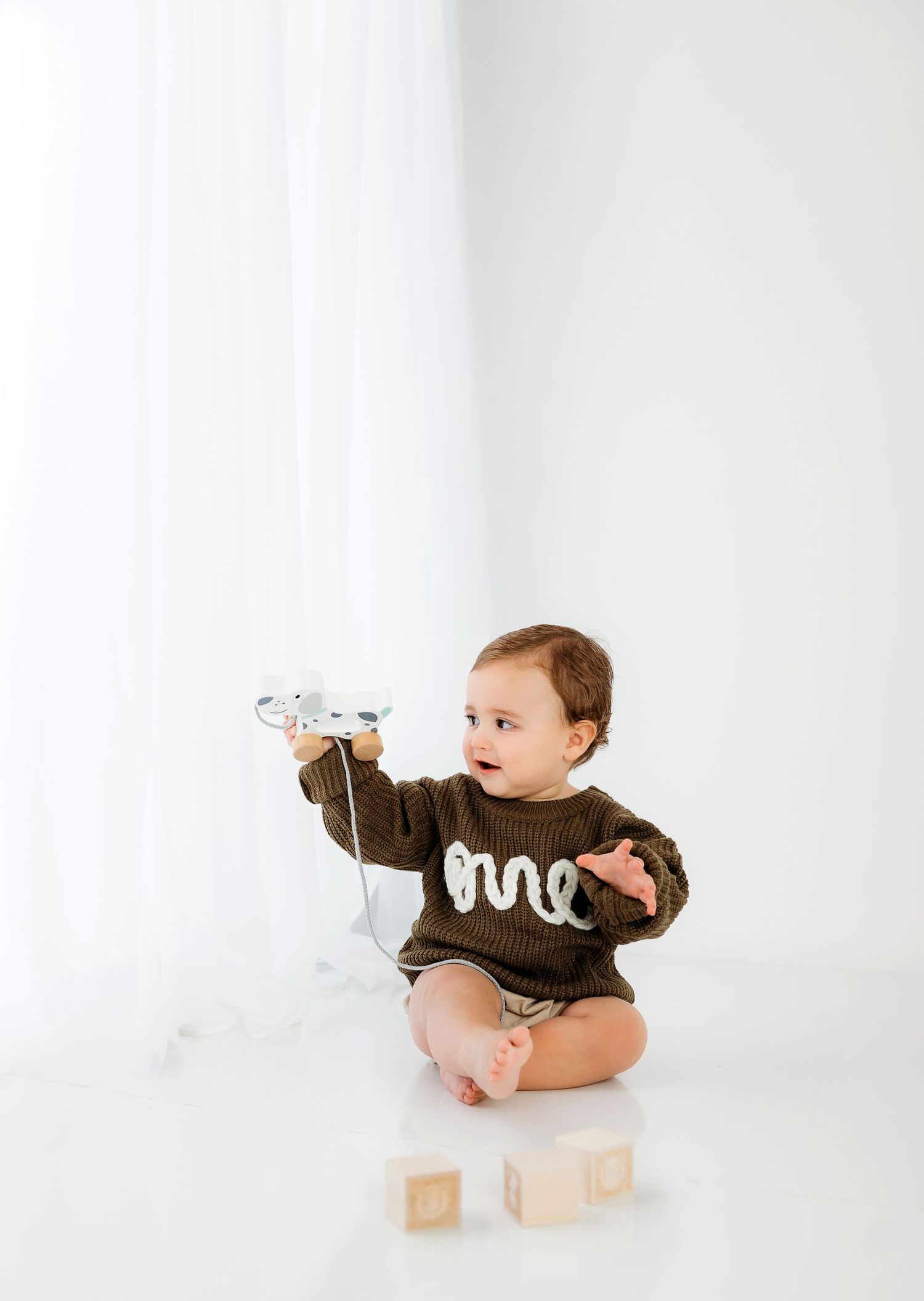 Baby in brown onesie sits on white floor with wooden blocks in minimalist studio setting.