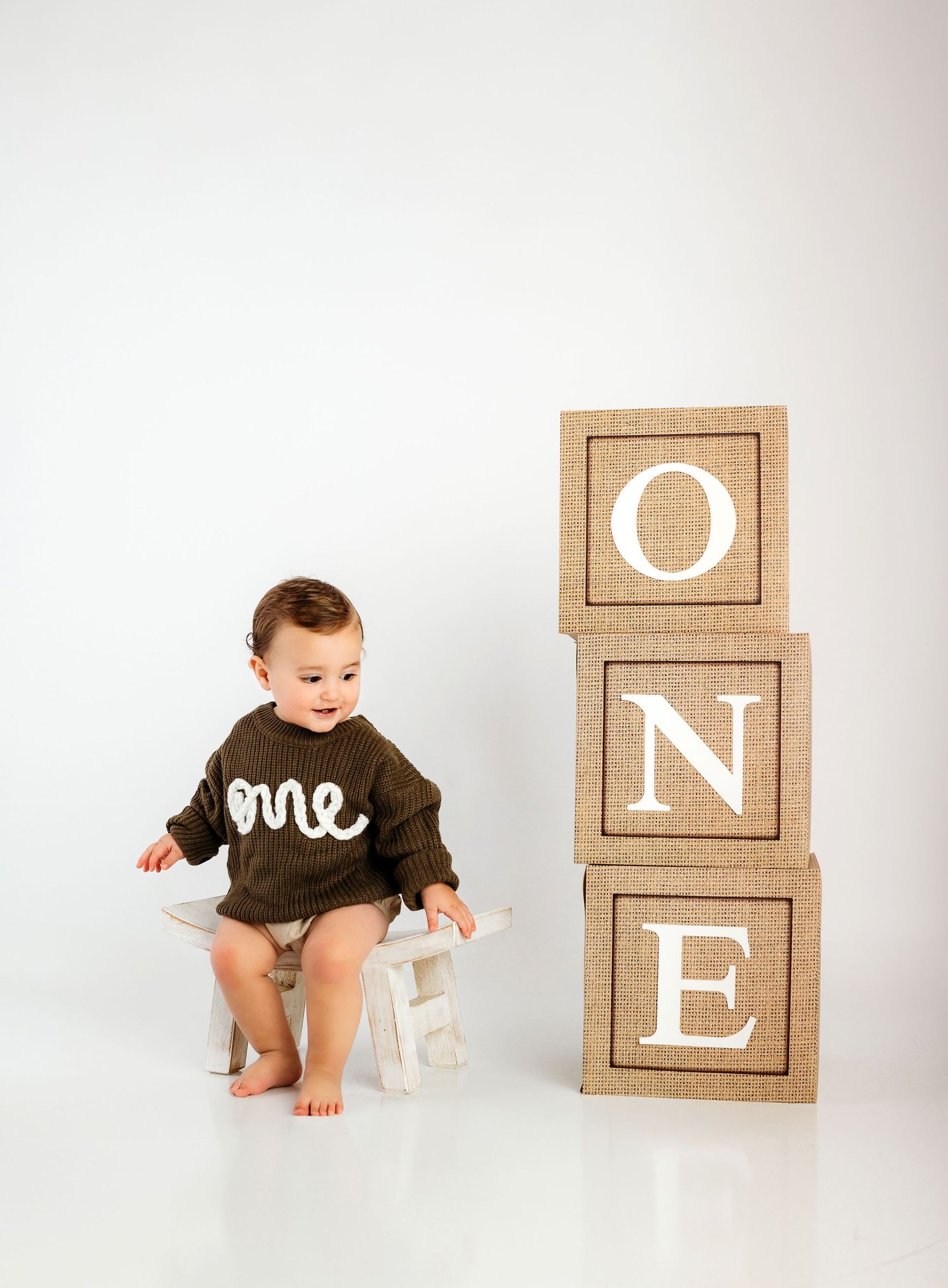 Small child in brown sweater sits on white stool next to large decorative wooden blocks spelling ONE against white backdrop.