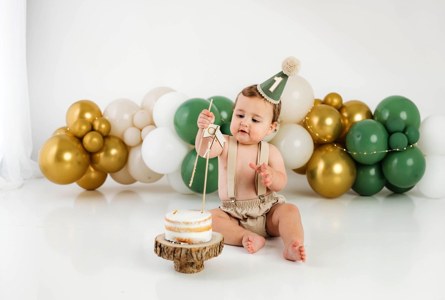 Baby in white dress poses with green and gold balloon arch during milestone photo session.