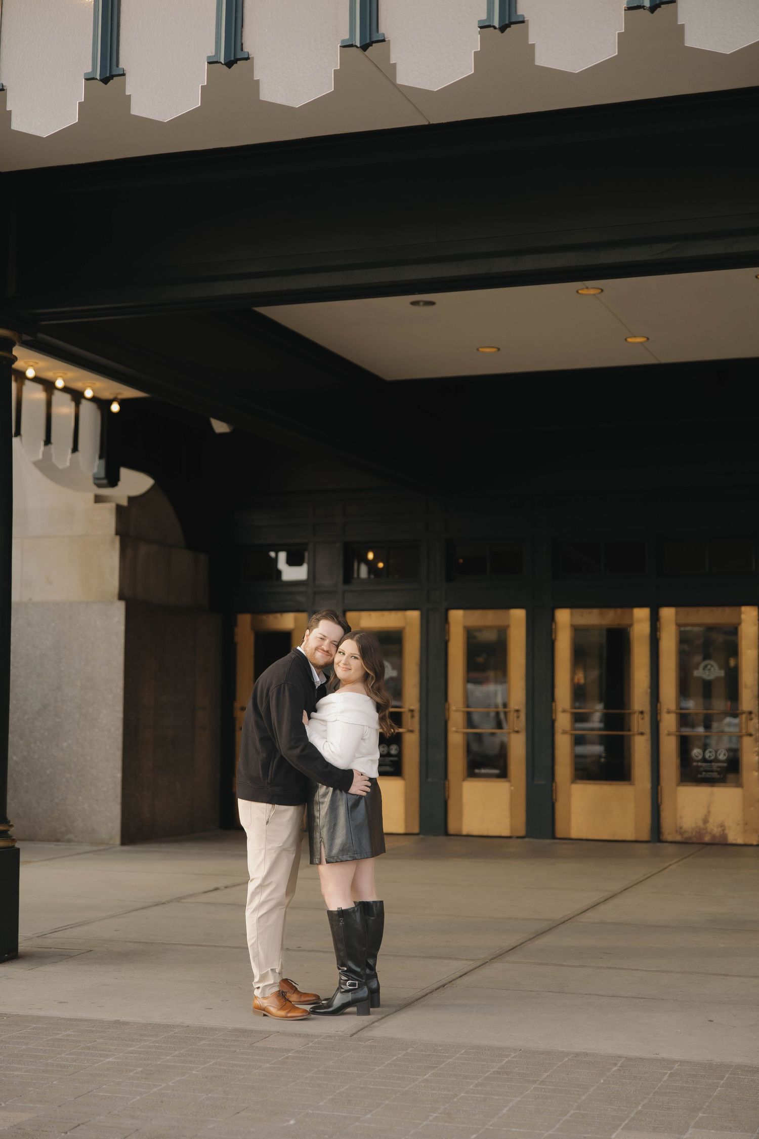 A couple embraces in front of a modern building with large windows and columns during a romantic moment.