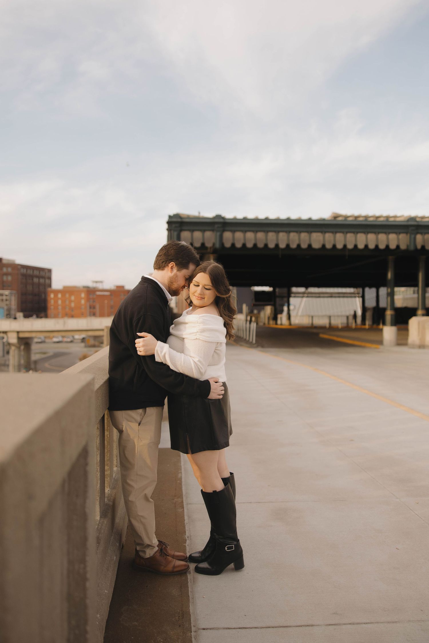 A romantic couple embraces on a concrete walkway with an industrial building and overpass in the background.