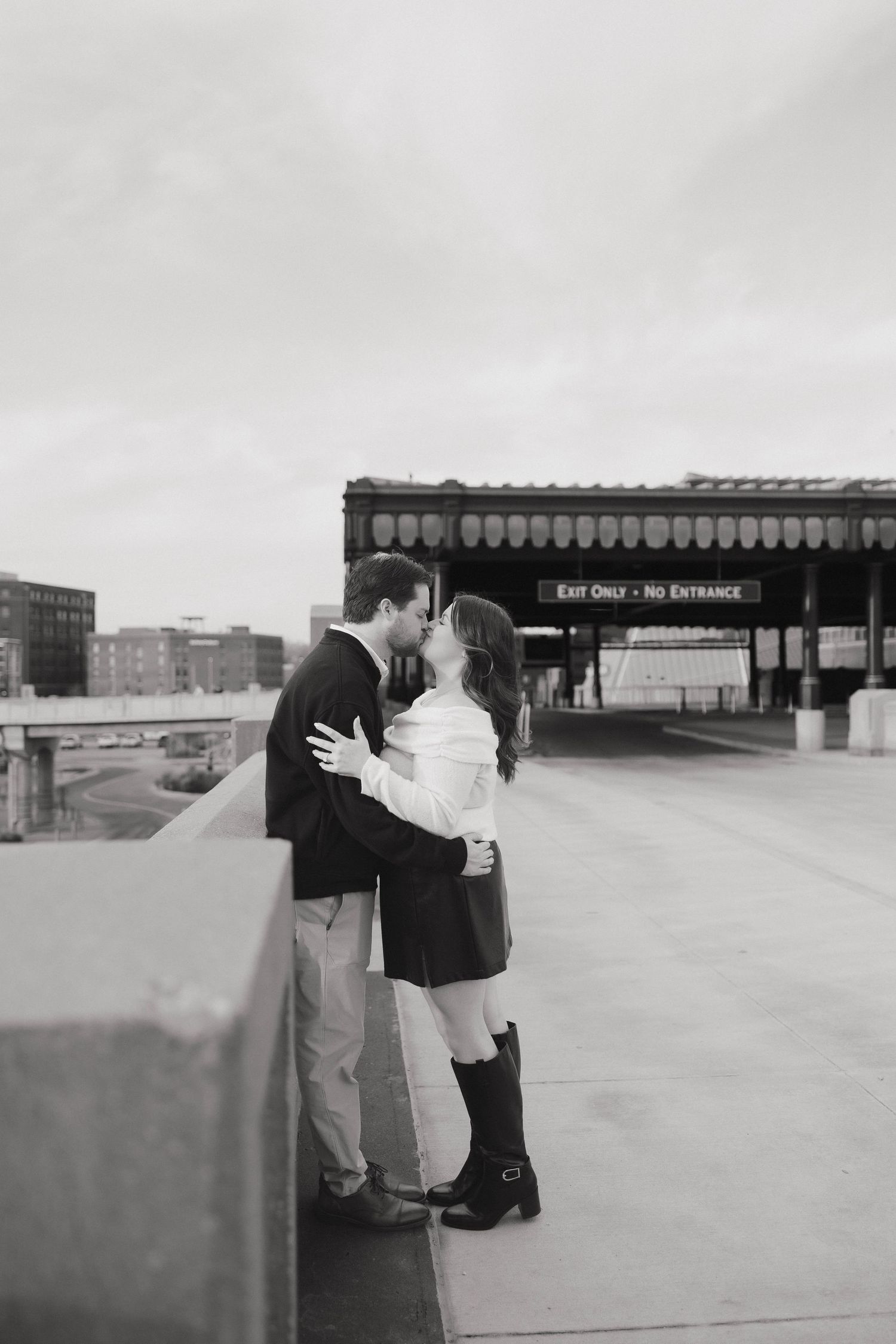A romantic black and white photo of a couple sharing a kiss on an urban rooftop with city buildings in the background.