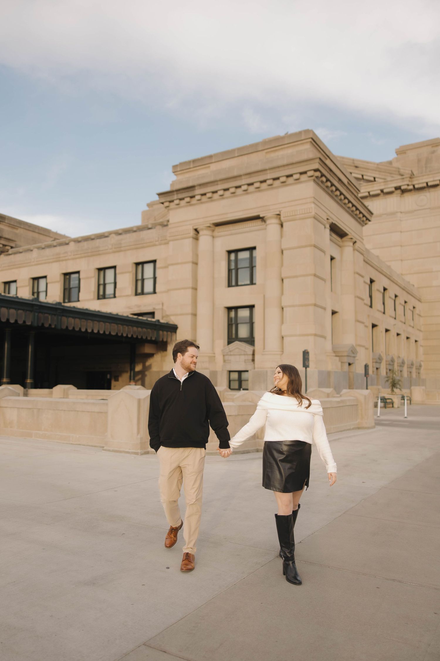 A couple walks together outside a grand beige stone building on an overcast day.