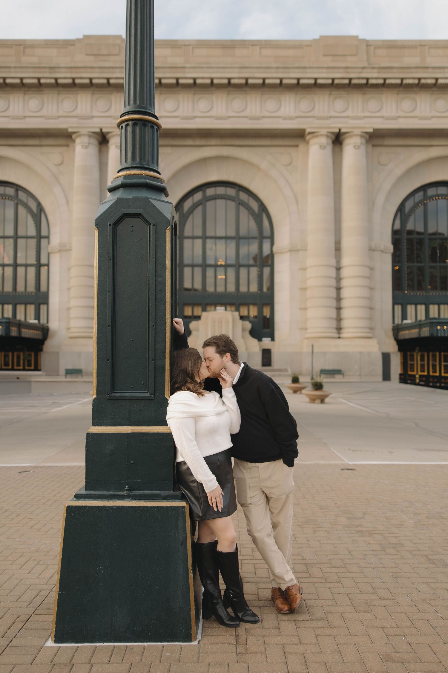 A couple shares an intimate moment by a vintage lamp post outside a historic building with arched windows.