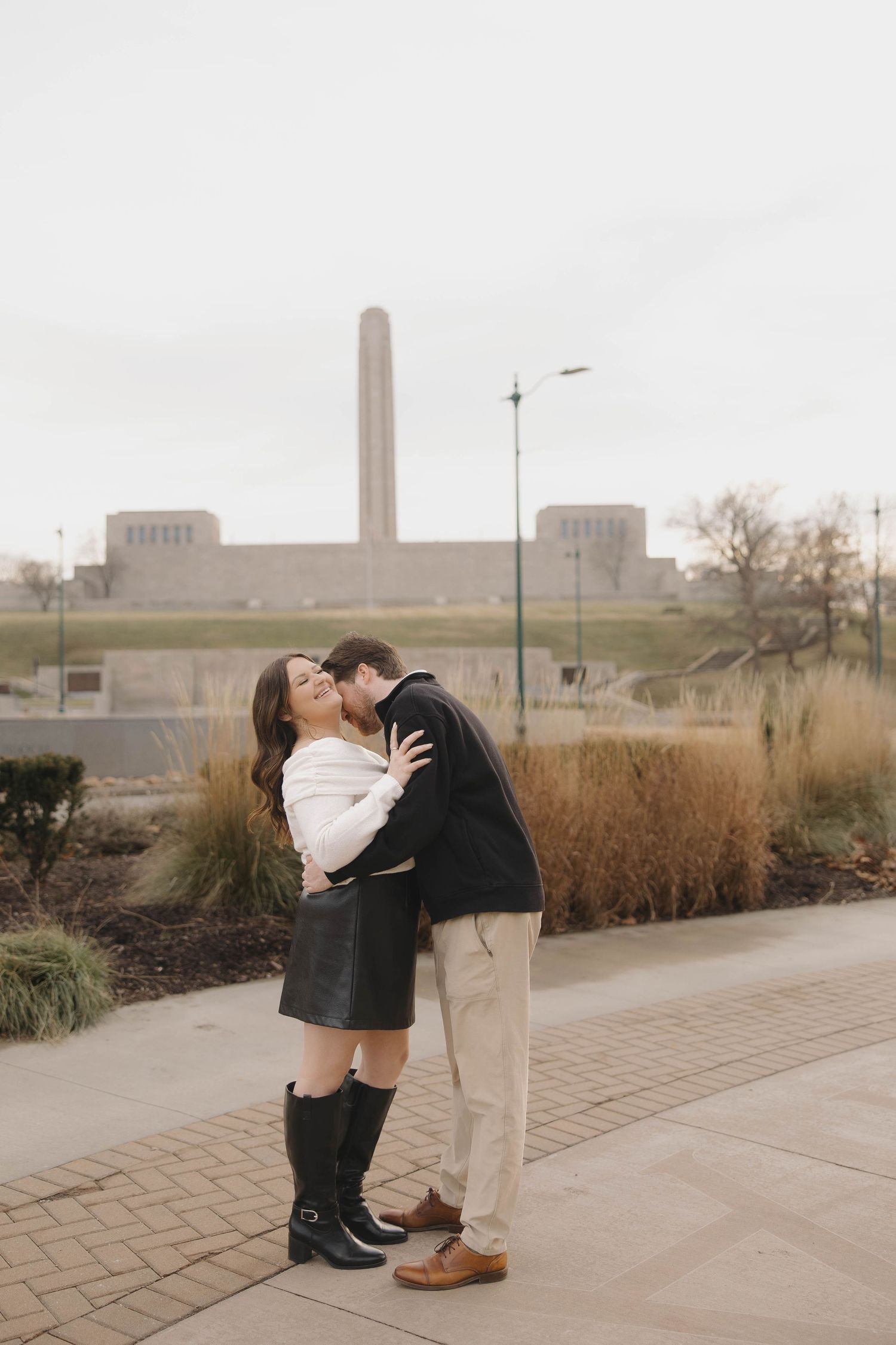 A couple shares an intimate moment in front of a tall monument on a cloudy day.