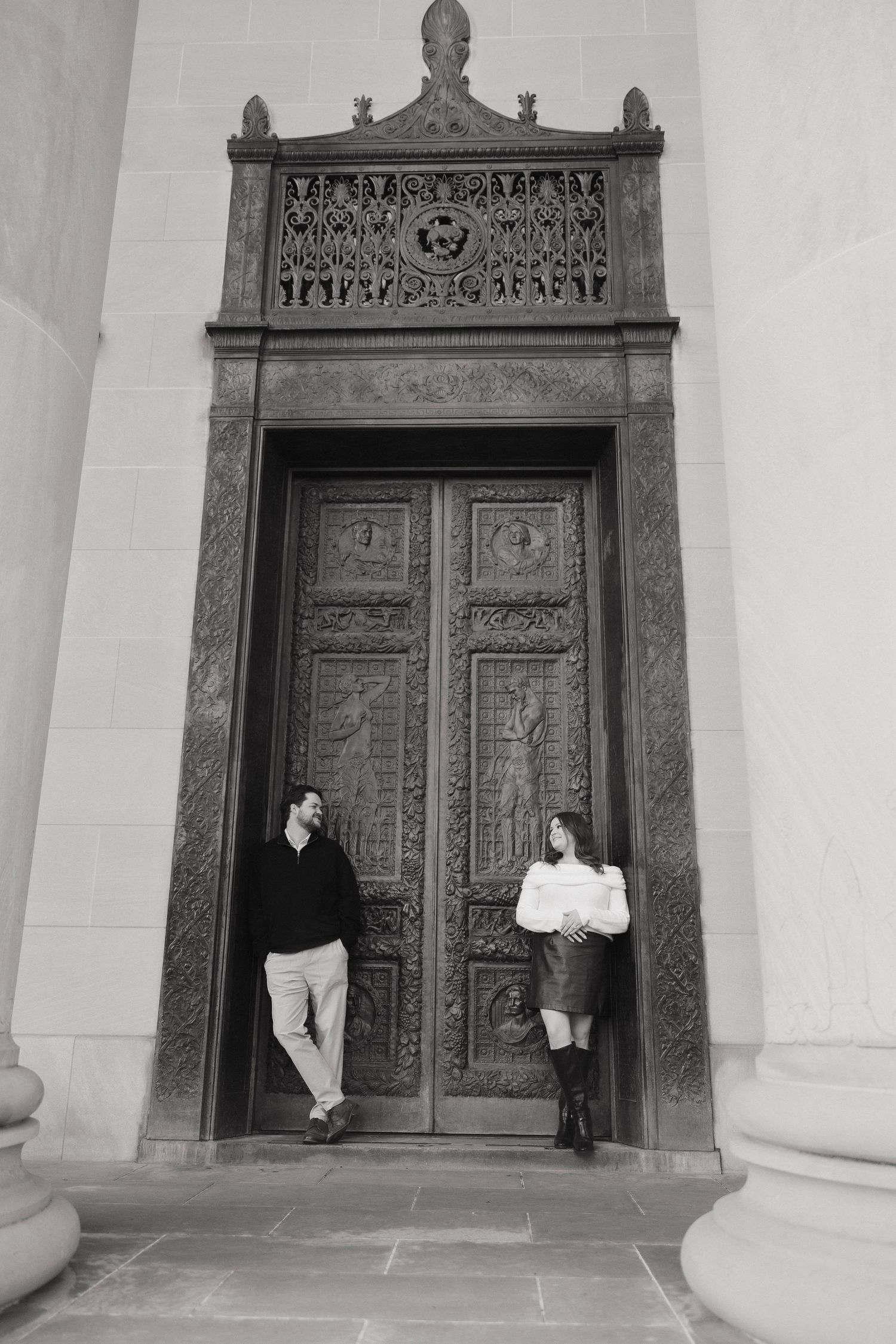 A couple poses playfully against an ornate carved wooden door in a black and white architectural photo.