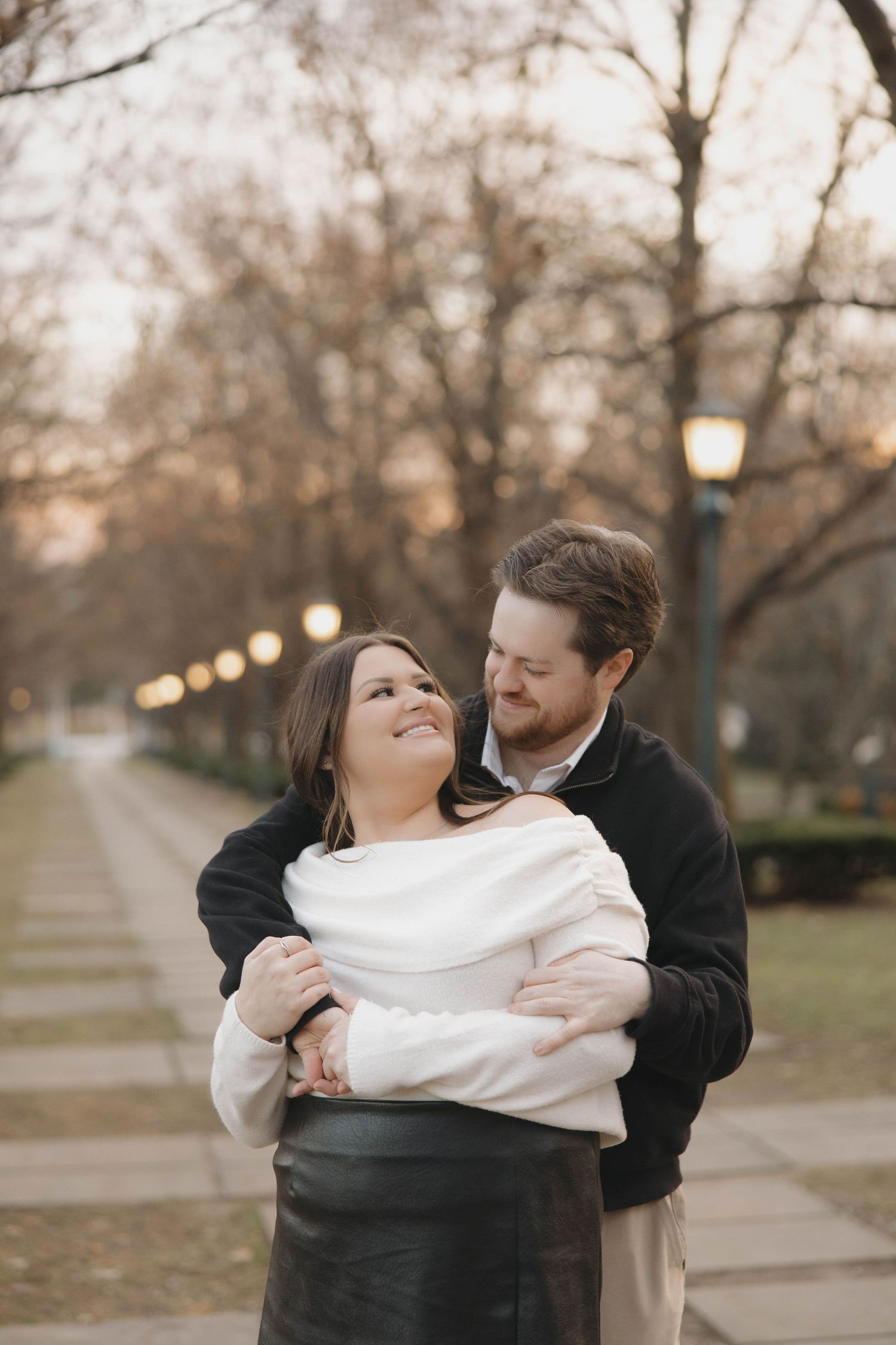 A romantic couple embraces outdoors with glowing lanterns and trees in the background at dusk.