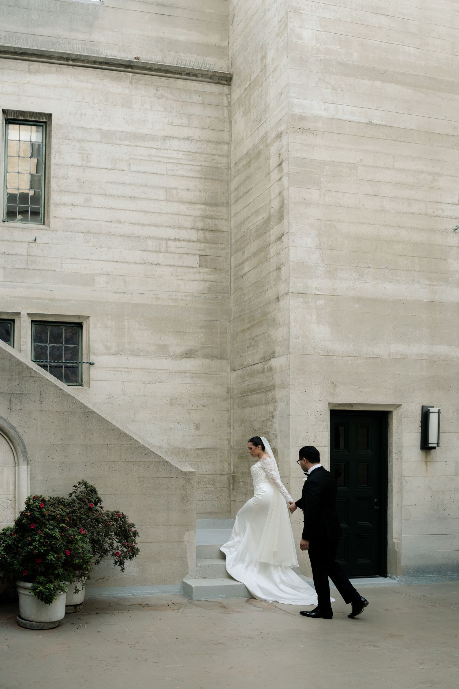 A couple in wedding attire ascend stone steps beside a white building with architectural details and potted plants.