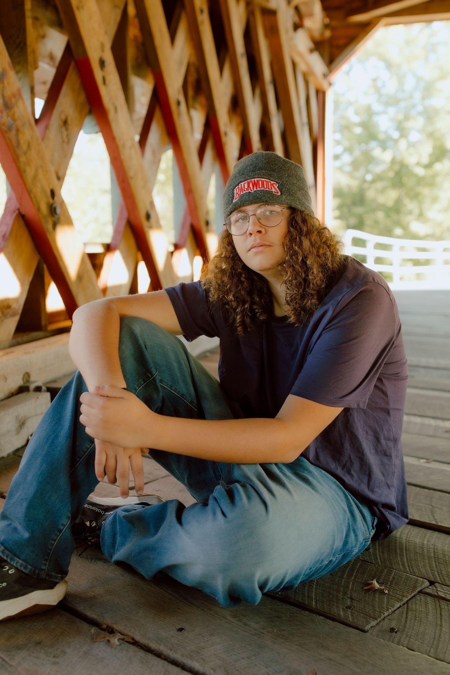 Young person with curly hair sitting under a covered wooden bridge wearing a beanie and blue jeans.