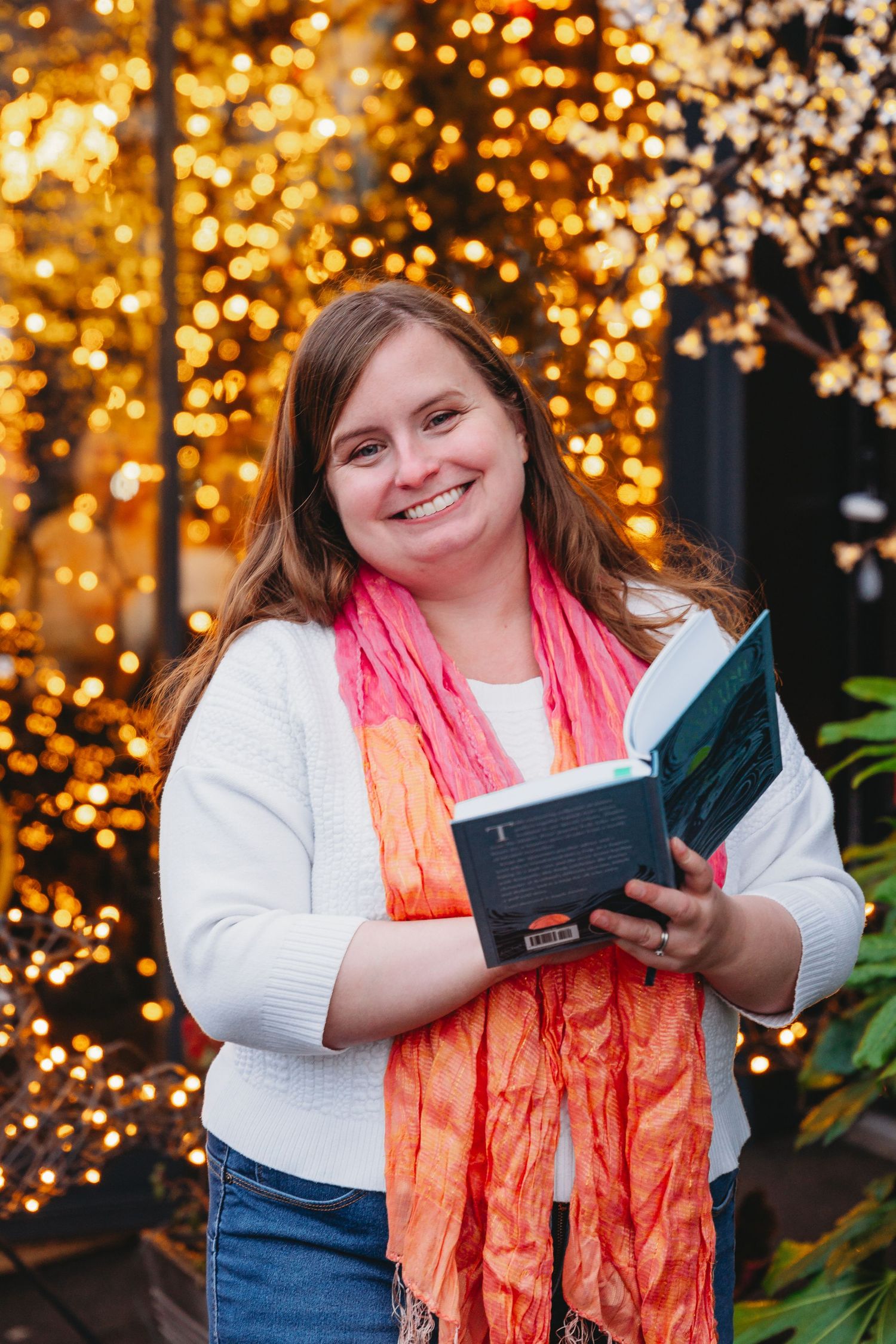 A person in a white sweater and coral scarf holds a book while standing in front of twinkling holiday lights.