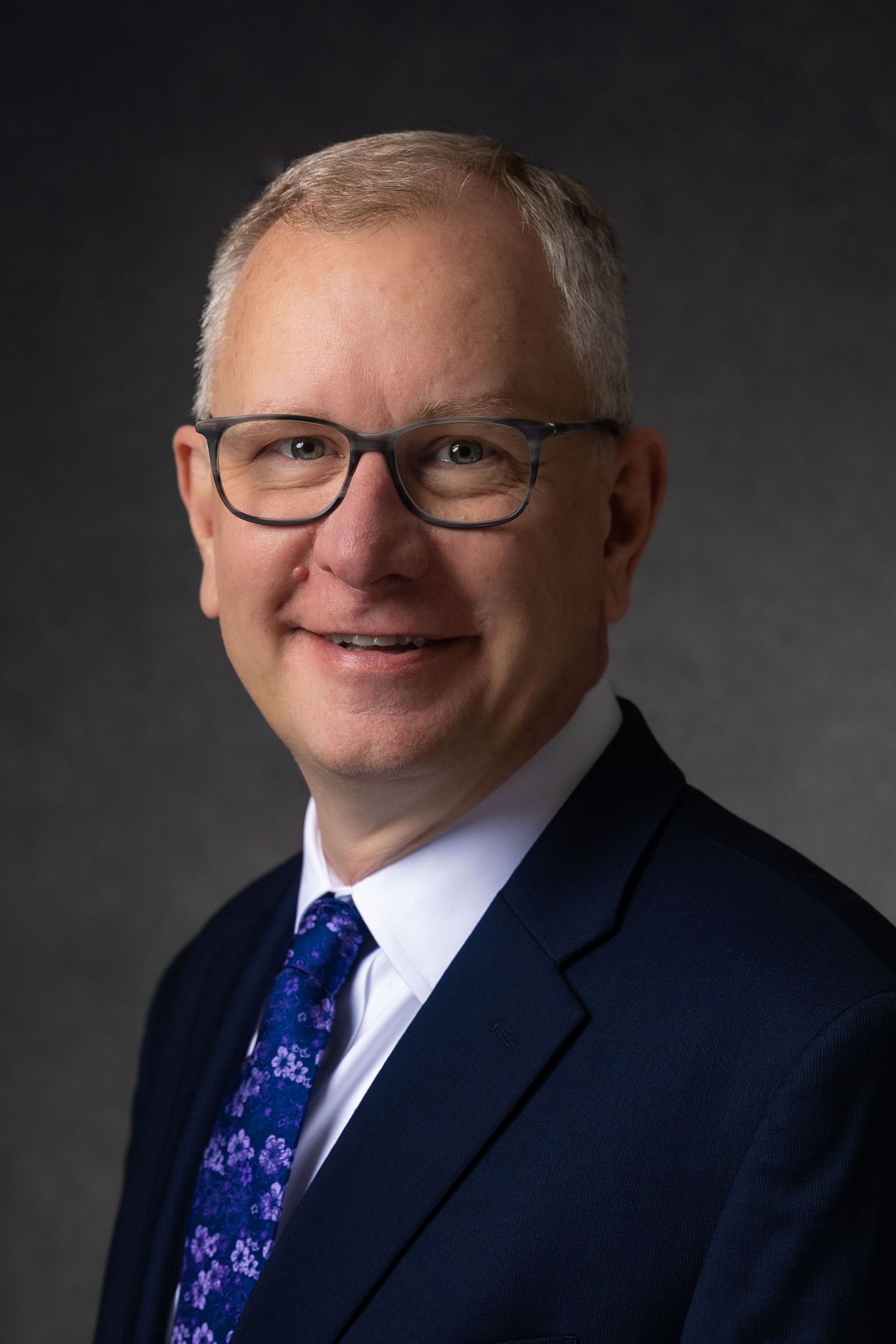 Professional headshot of a middle-aged man with glasses, gray hair, wearing a navy suit and floral tie.