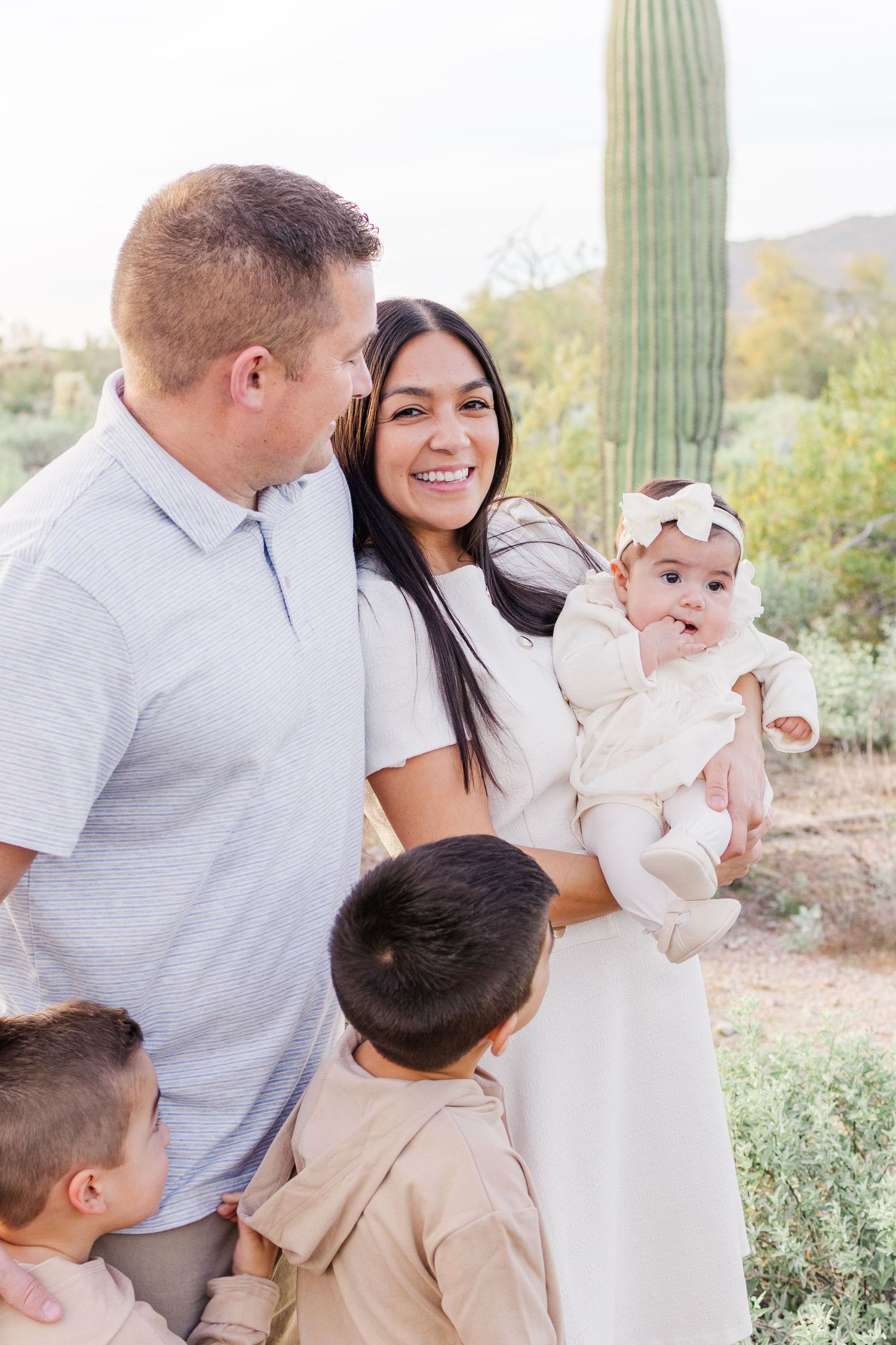 mom smiling at camera holding infant daughter while cuddled up with her husband and two boys next to a cactus in the desert
