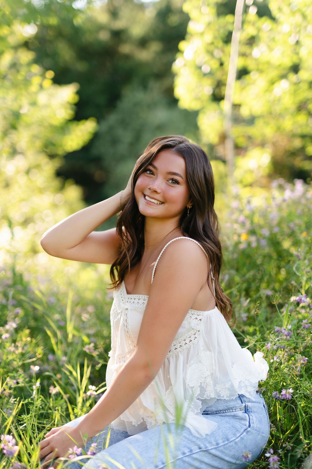 A person in a white lace top and jeans sits in a sunny meadow with wildflowers and greenery.
