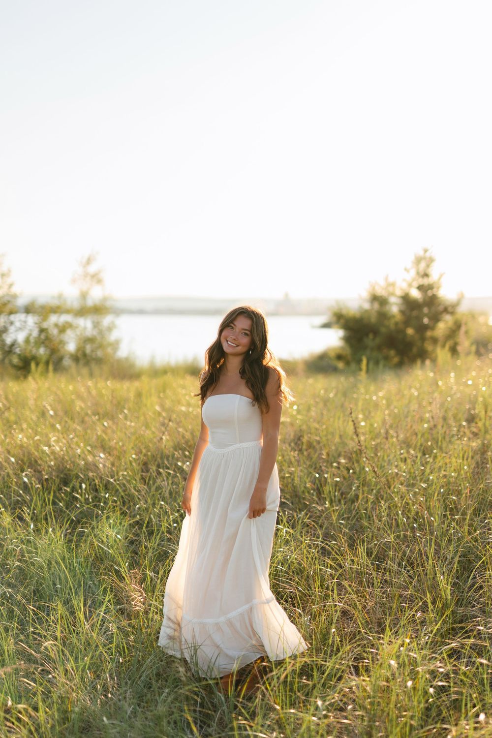 A woman in a flowing white maxi dress stands in a sunlit field near a lake during golden hour.