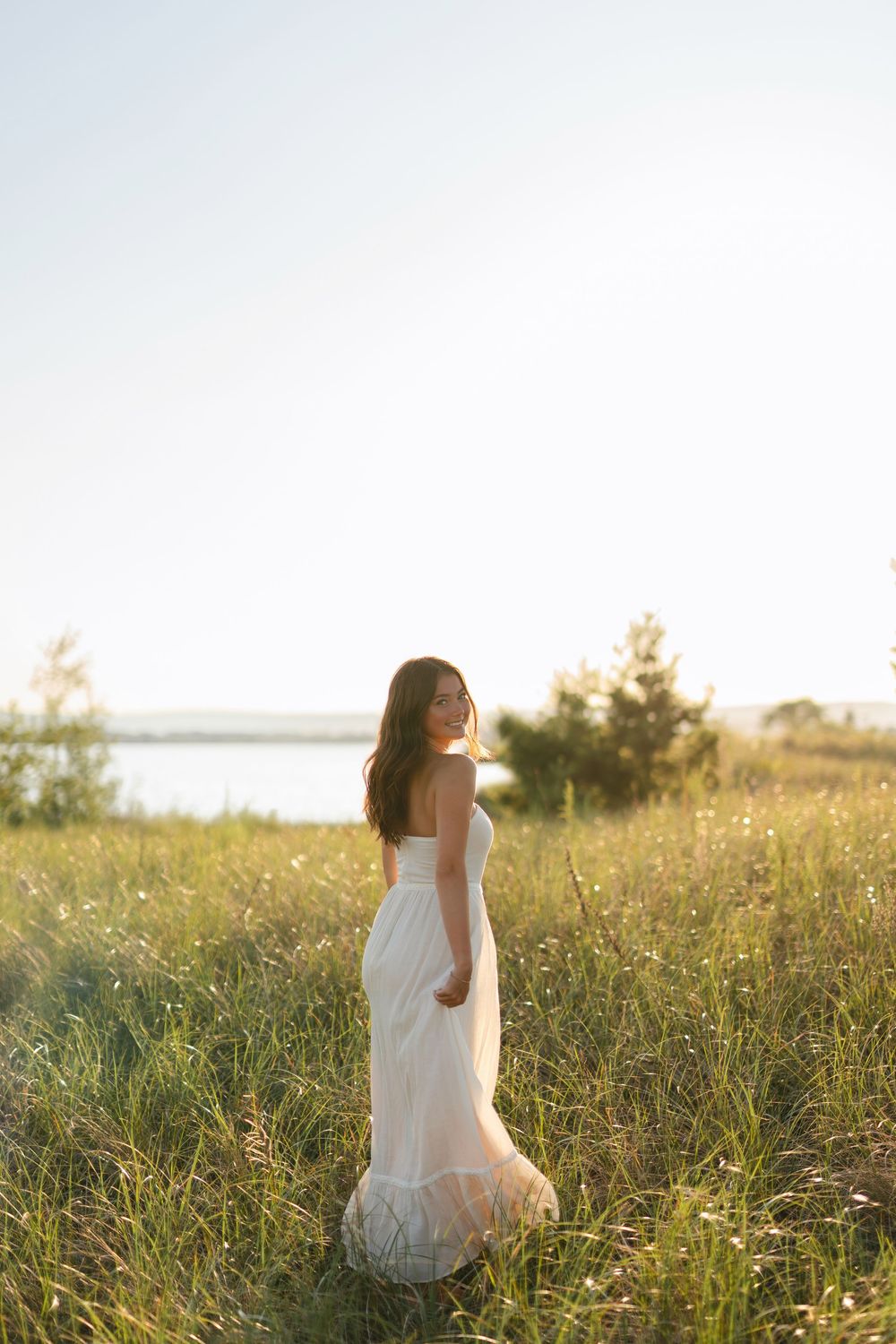 A woman in a flowing white dress stands in a sunlit meadow near water during golden hour.