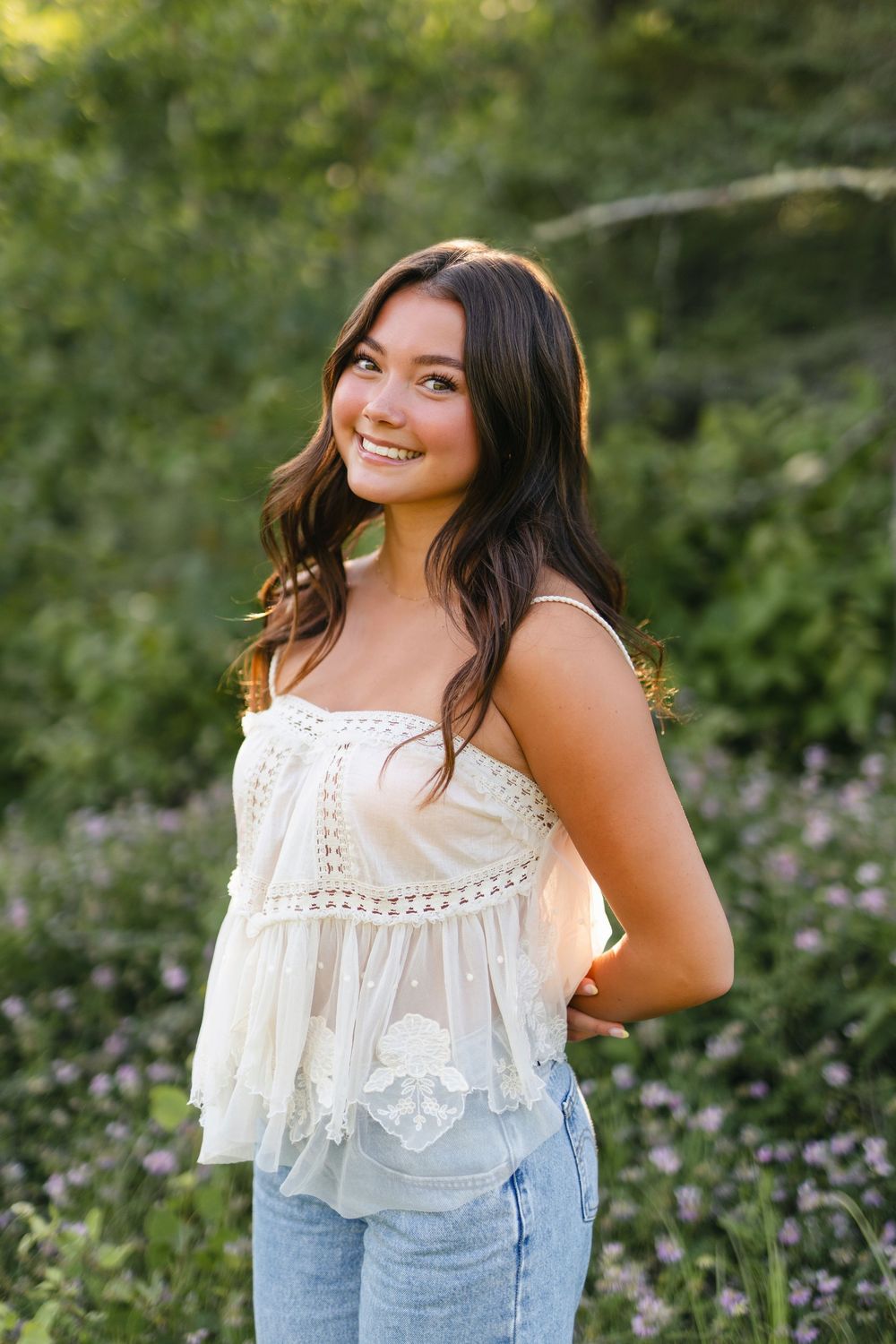 A white lace crochet crop top paired with denim is photographed in a garden setting with purple wildflowers.