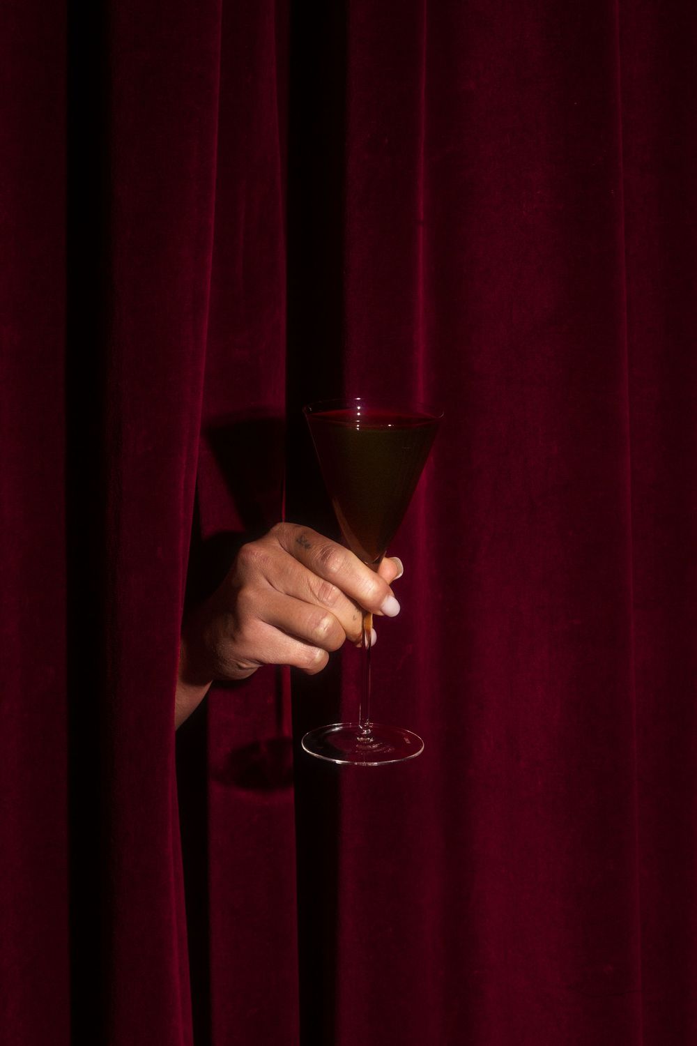 Hand holding a cocktail glass against a dark red curtain backdrop.