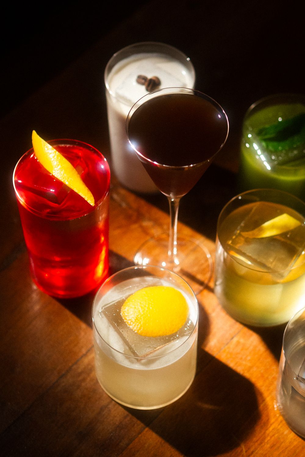 Colorful cocktails with garnishes arranged on a wooden bar surface in dramatic lighting.