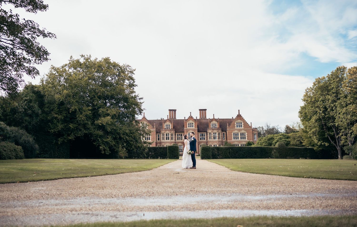 A panoramic view of a gravel path leading to historic brick buildings surrounded by green trees and manicured lawns.