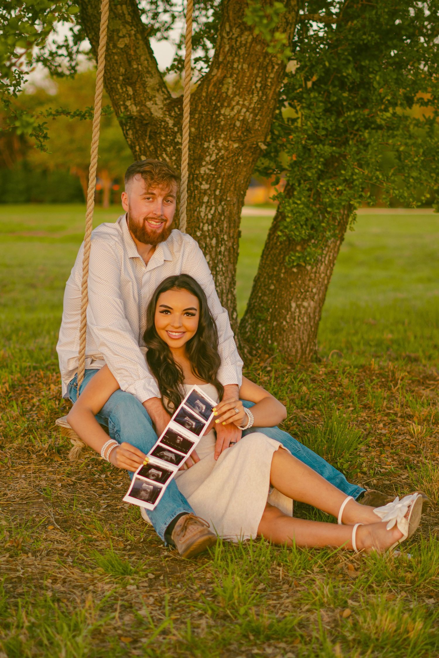 Couple sits on tree swing holding ultrasound photos in park setting.