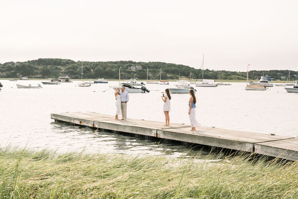People standing on a wooden dock overlooking a harbor with boats and grassy shoreline on a summer day.