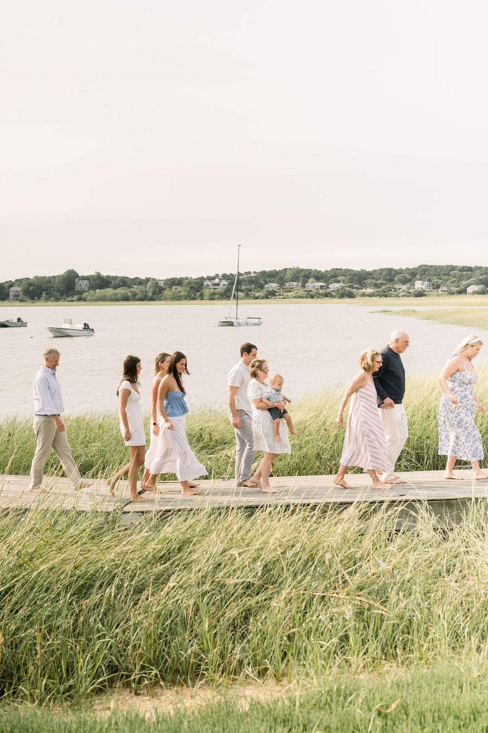 A panoramic sequence shows people walking through tall coastal grass along a shoreline path on a sunny summer day.