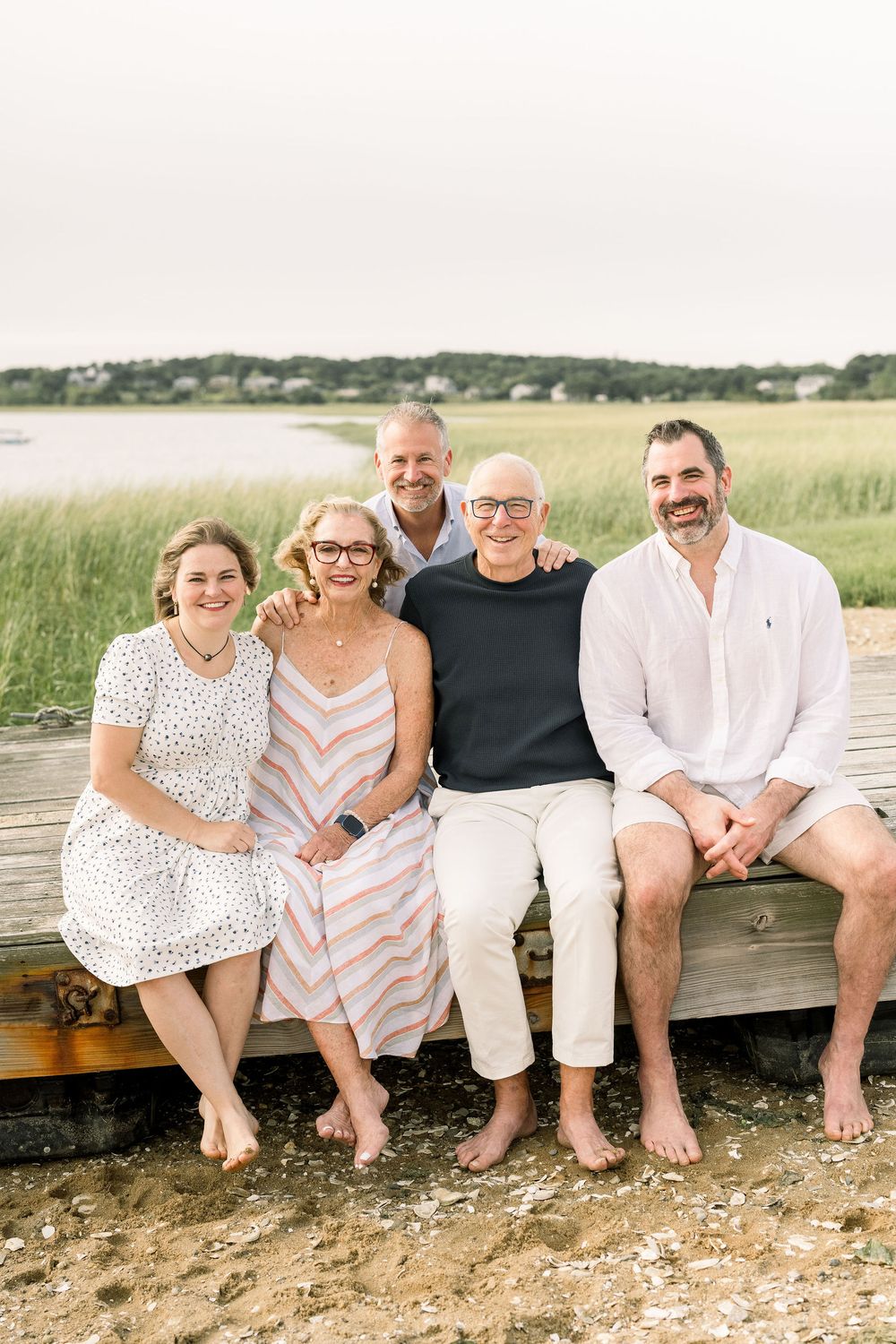 A group relaxes together on a wooden bench near a coastal marsh on a summer day.