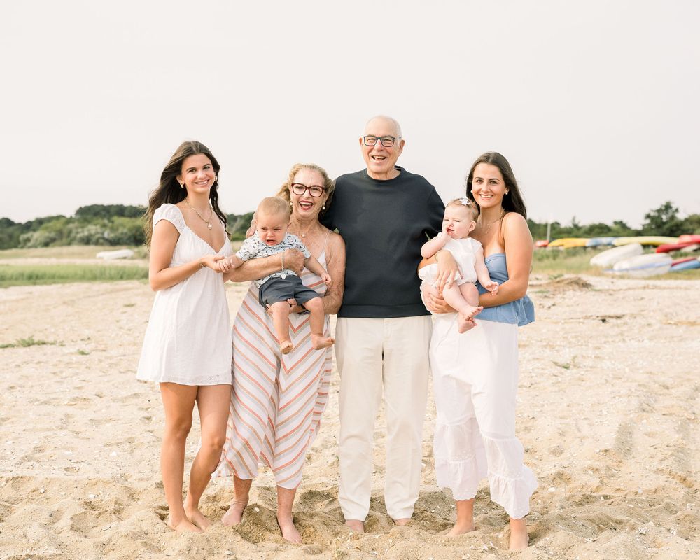 Family wearing coordinated white and beige outfits poses together on a sandy beach at sunset.