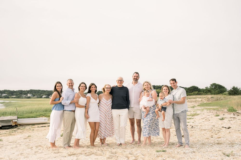 A group of people in coordinated white and beige outfits pose together for a family photo session on a sandy beach.