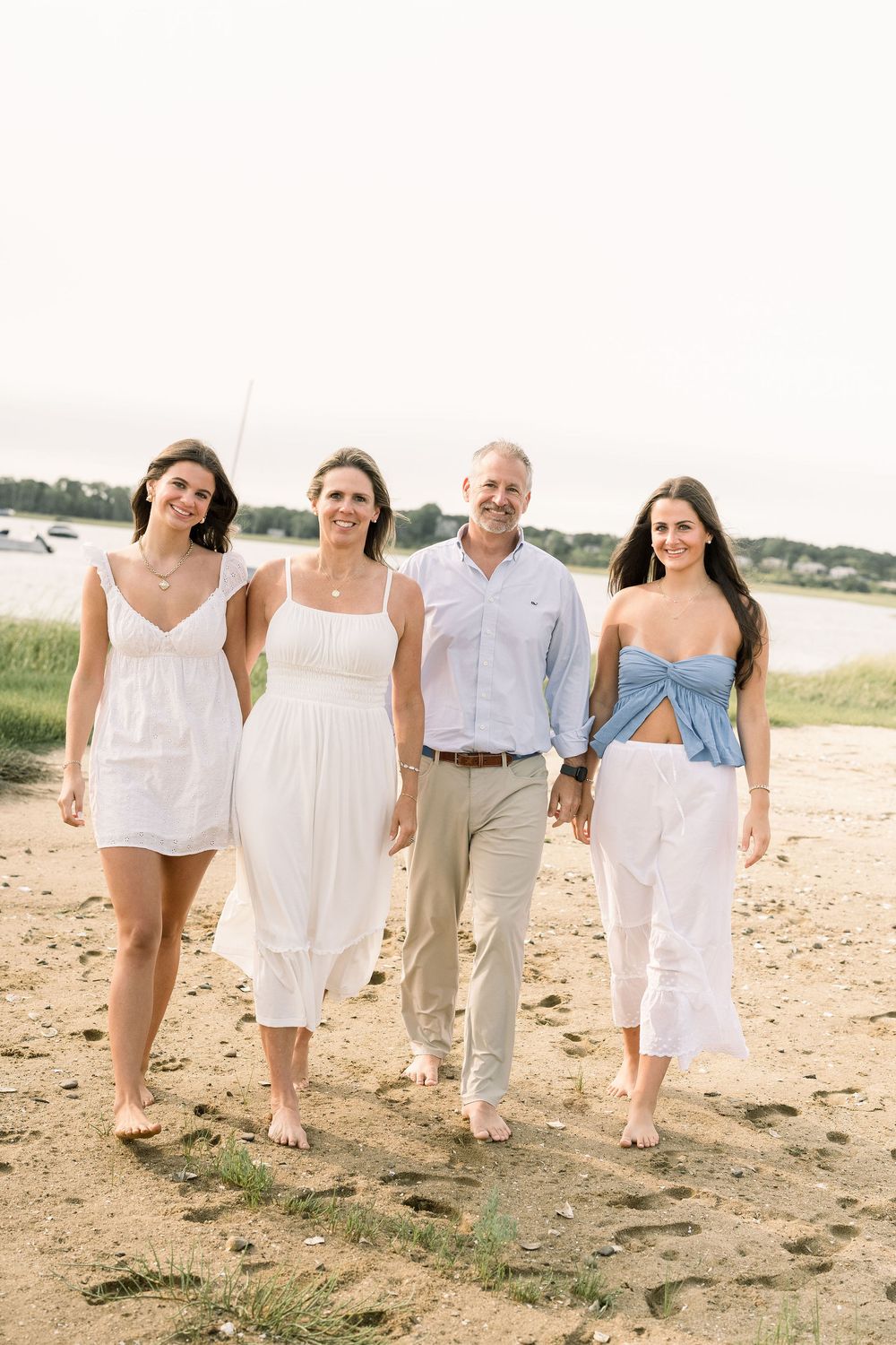 A group in white summer outfits walks together barefoot along a sandy beach on a sunny day.