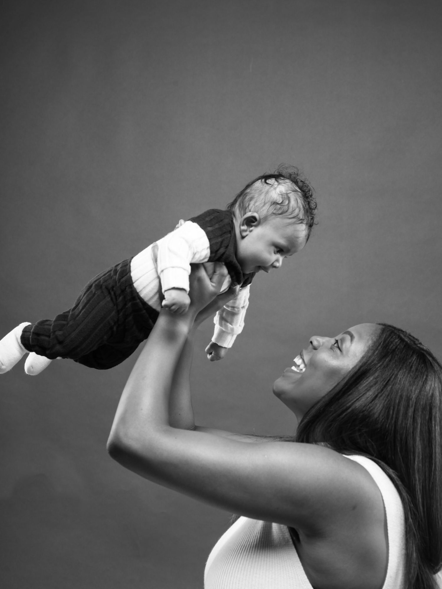 A tender black and white portrait shows a joyful parent lifting a baby up in the air against a dark background.