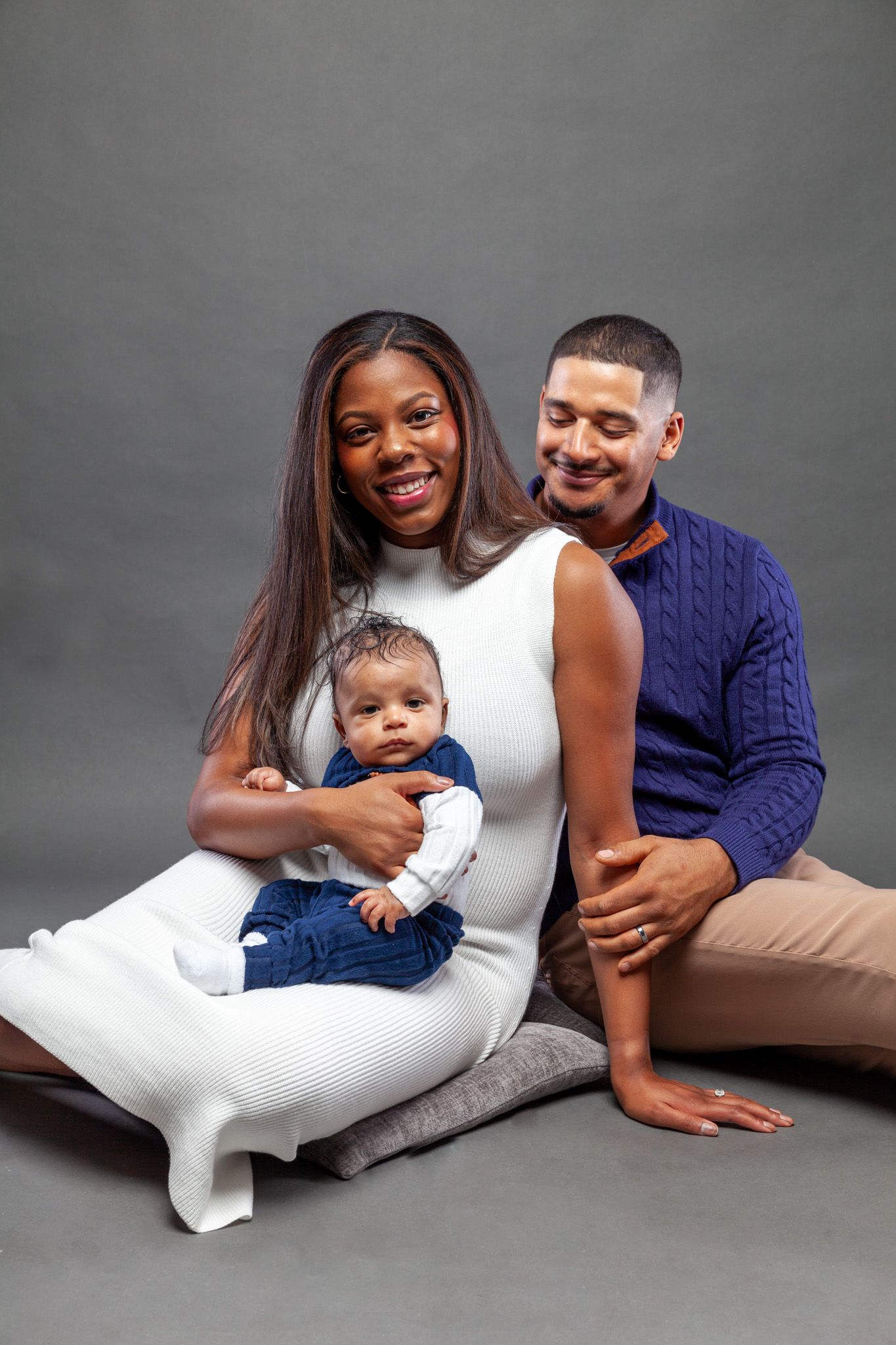 A family in coordinated white and navy outfits poses together lying down on a gray studio backdrop for portraits.