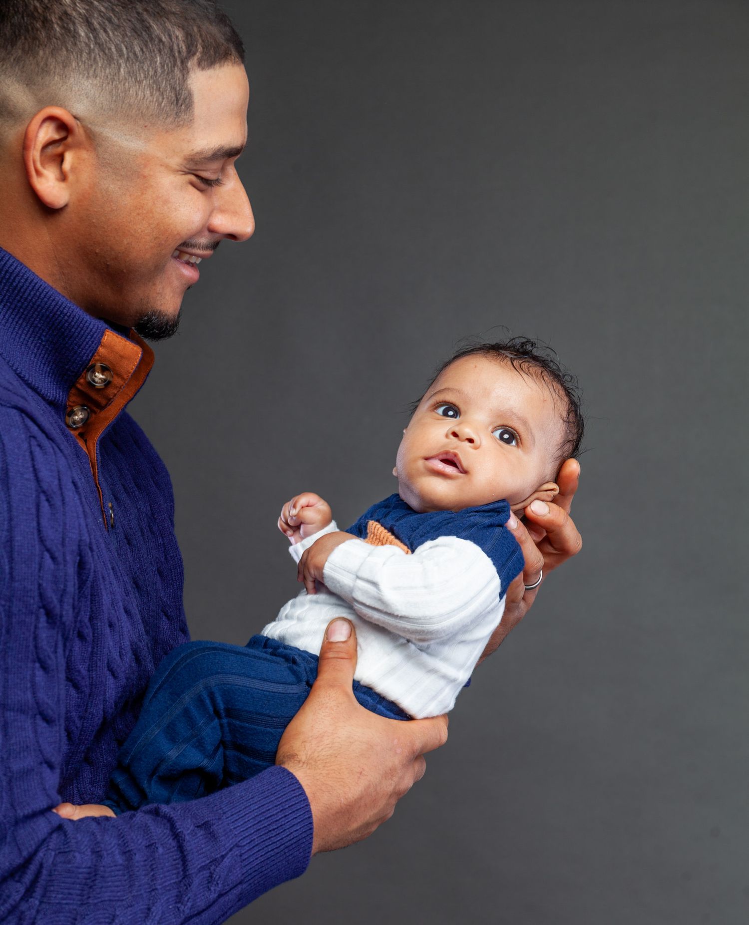 A parent in a blue sweater holds and plays with a smiling baby wearing white clothing against a dark background.