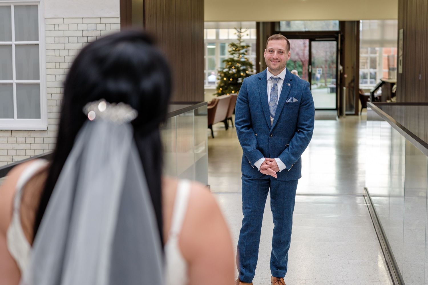 A groom in a navy blue suit sees his bride for the first time in a bright indoor hallway during their first look.