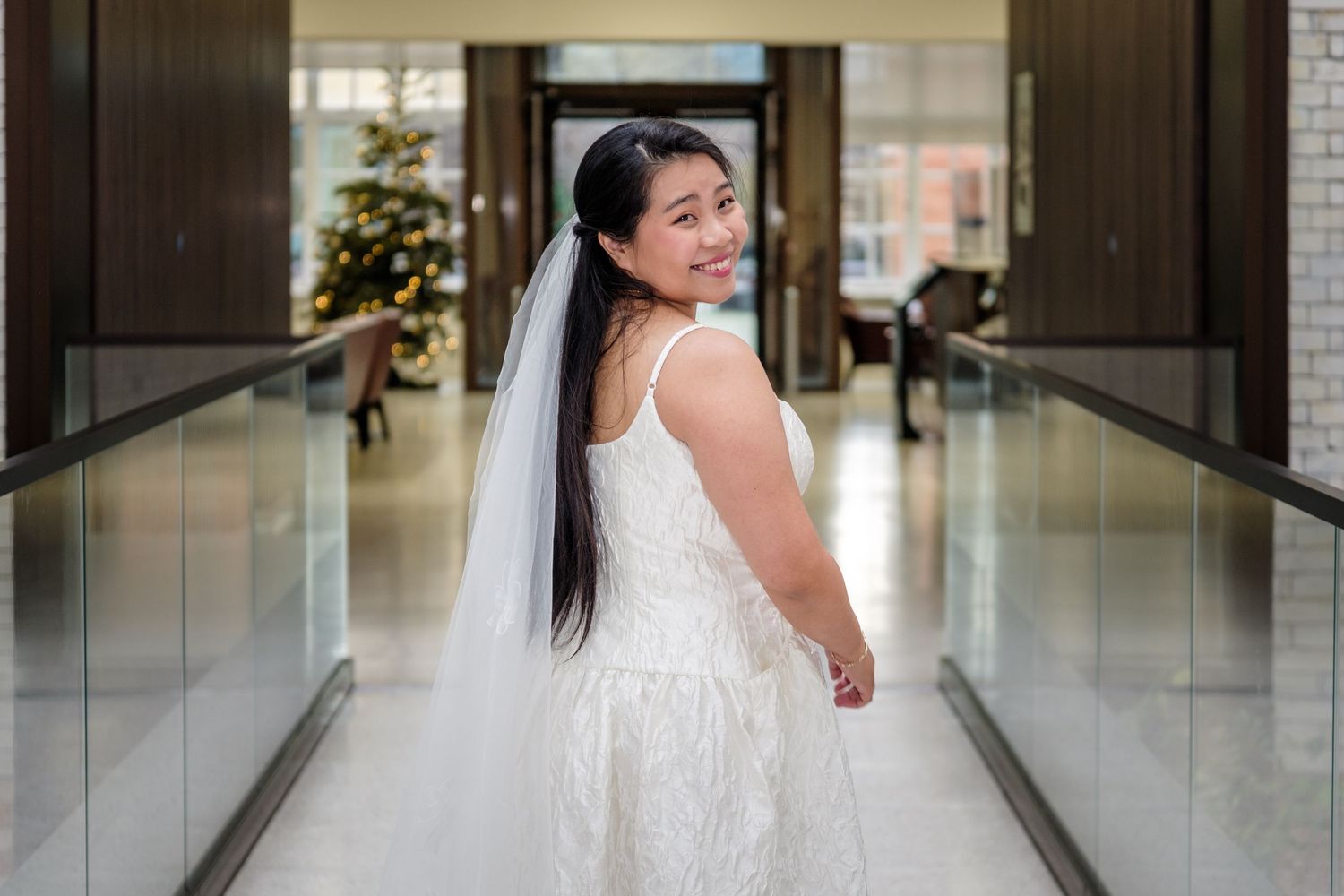 A bride in a white lace wedding dress poses elegantly in a bright indoor hallway.